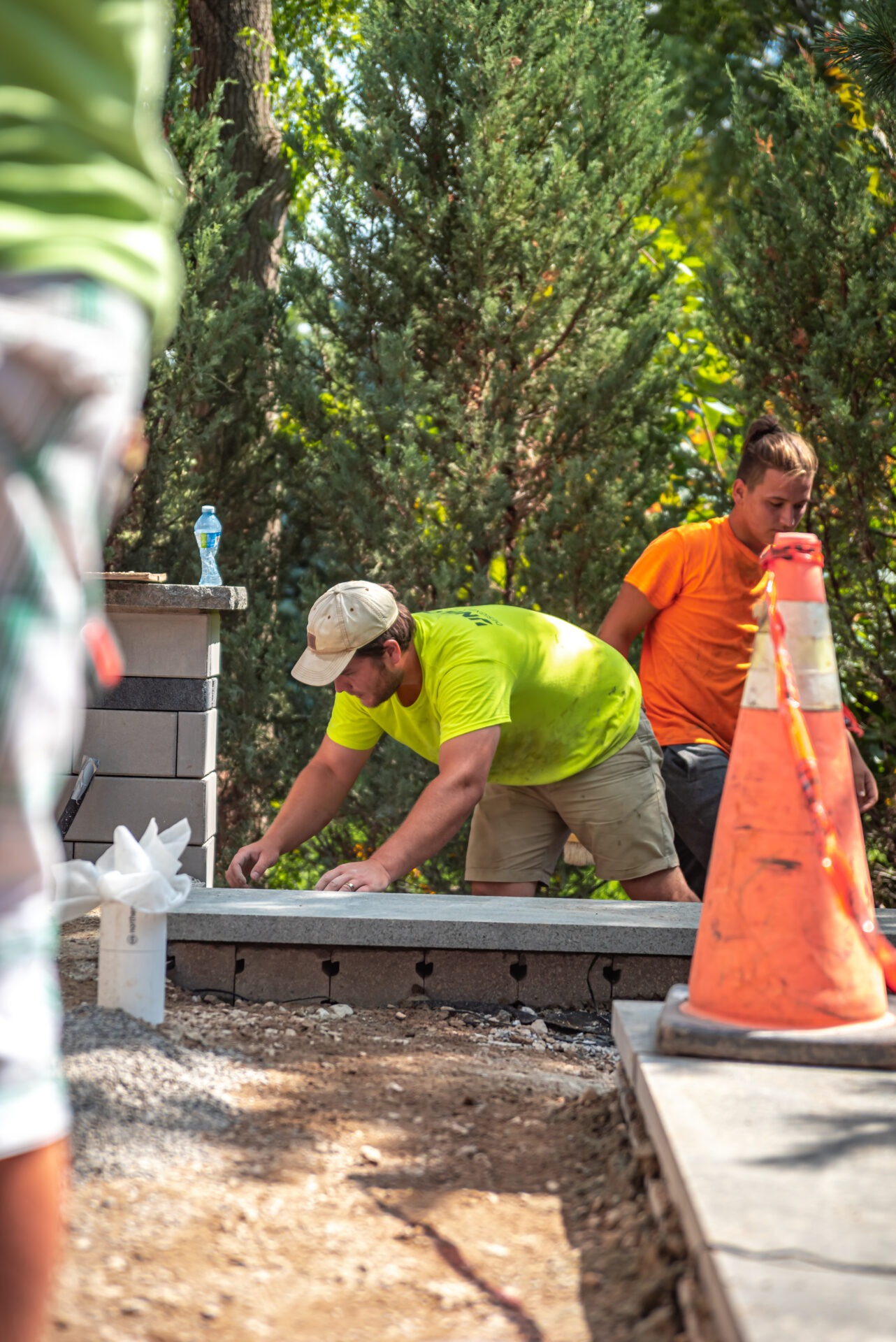 Two persons work outdoors near a construction site with cones and bricks. Trees surround them, creating a lush background.