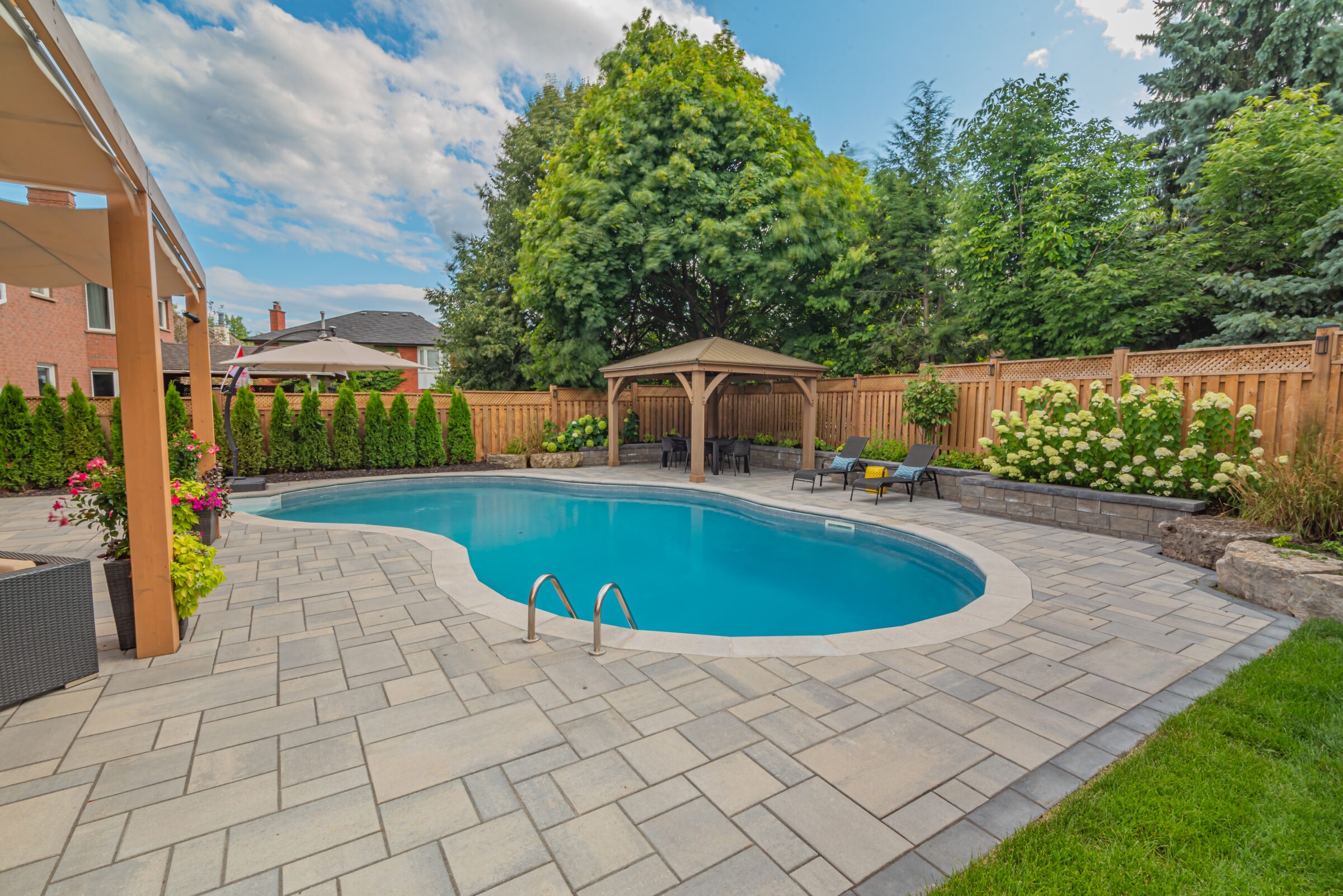 Inviting backyard pool surrounded by a wooden fence, lush greenery, and patio furniture under a gazebo. Clear sky with scattered clouds.