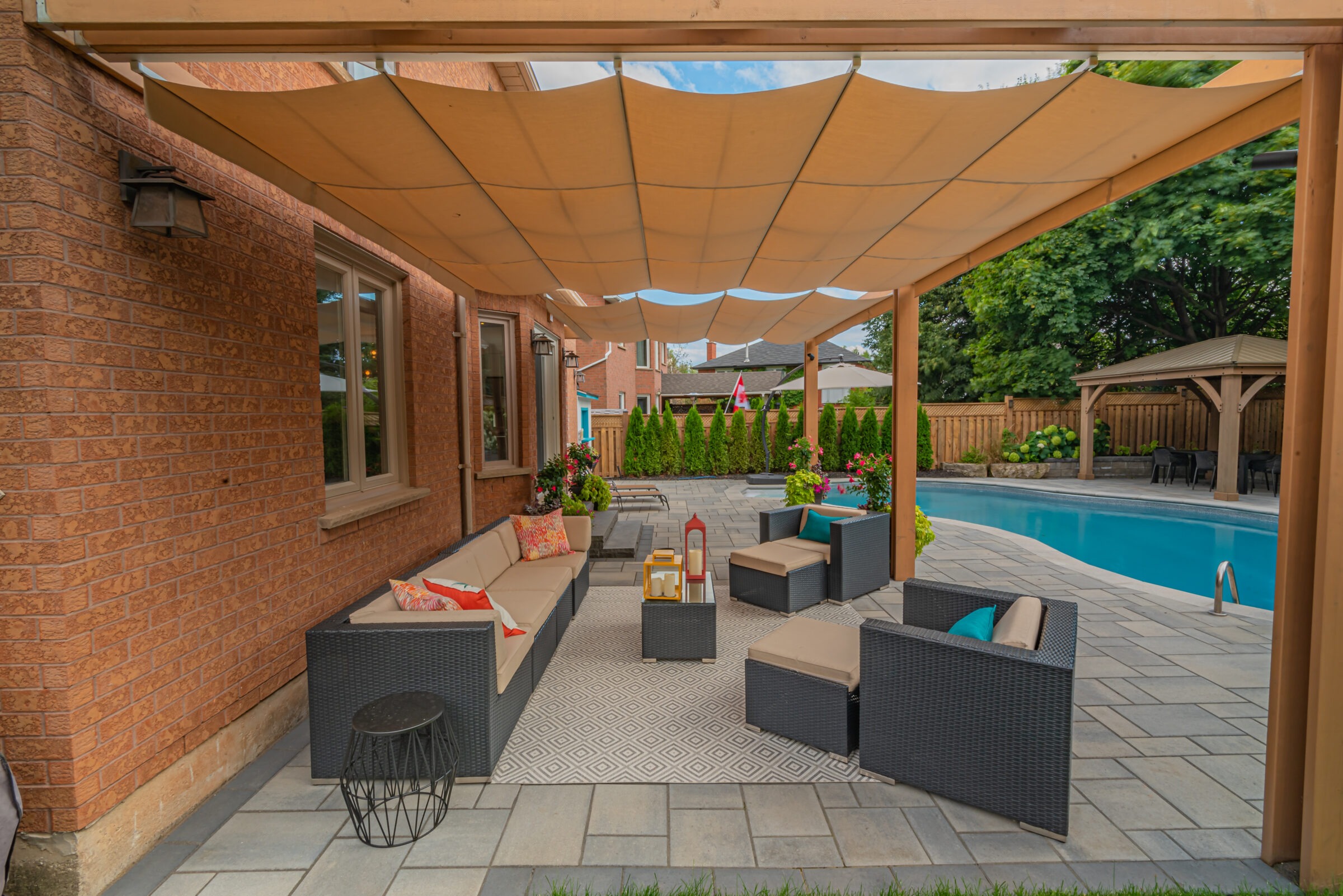 Patio with wicker furniture under a pergola by a backyard pool. Colorful cushions and plants create a cozy, inviting outdoor space.