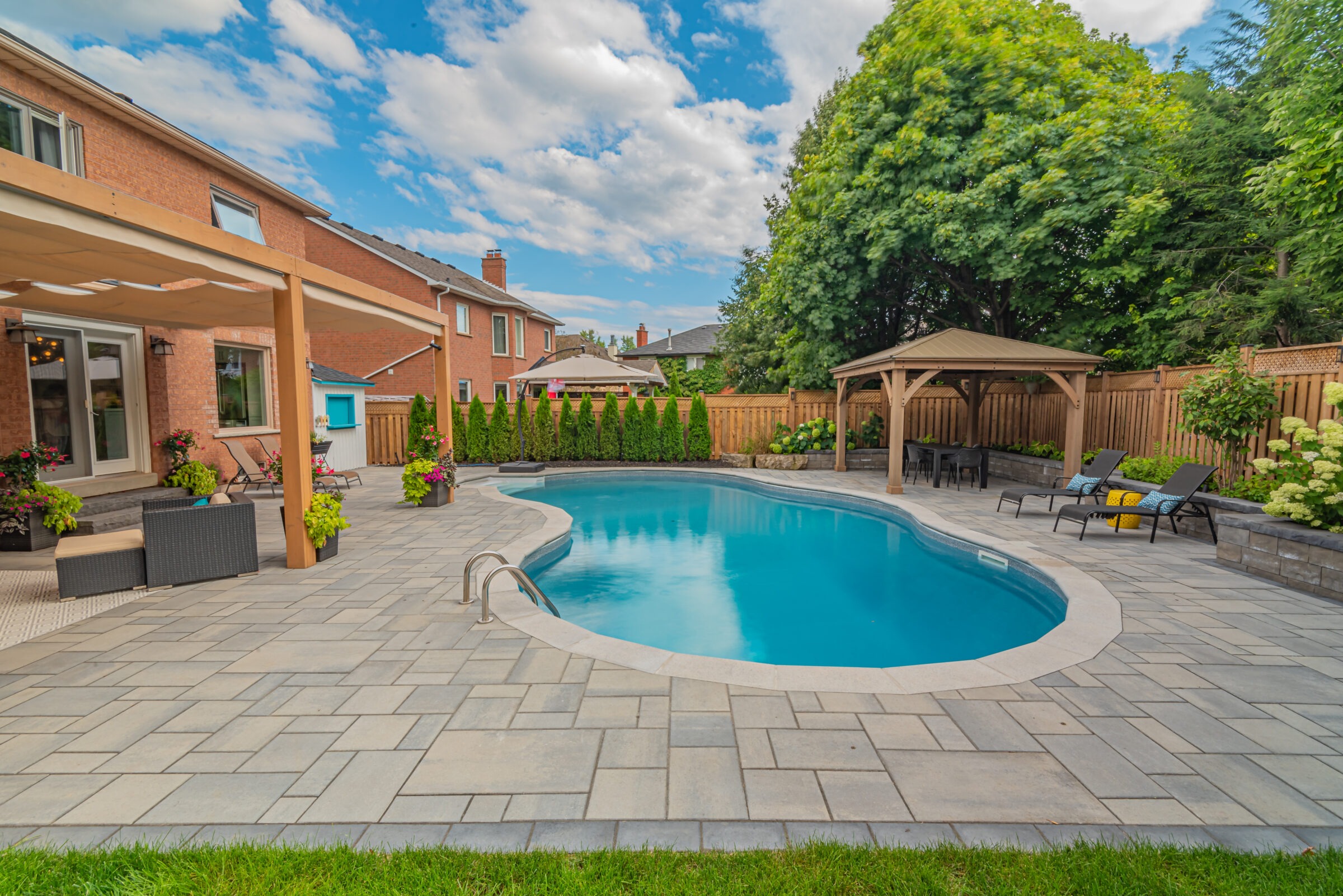 A backyard features a kidney-shaped pool, lounge chairs, gazebo, and lush landscaping, bordered by a wooden fence and adjacent to a brick house.