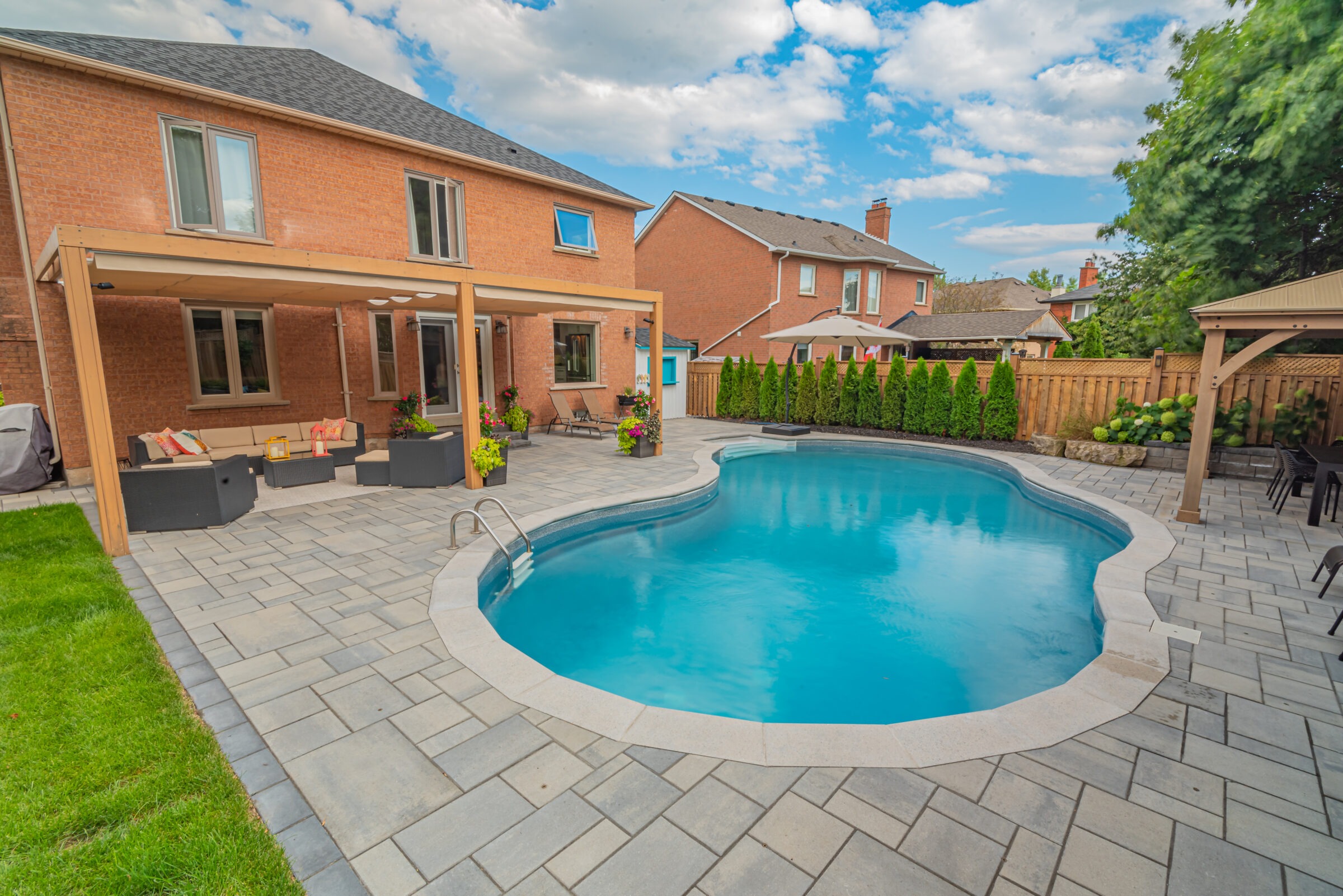 A backyard featuring a pool, patio with seating, and brick house under a partly cloudy sky. Peaceful residential setting.