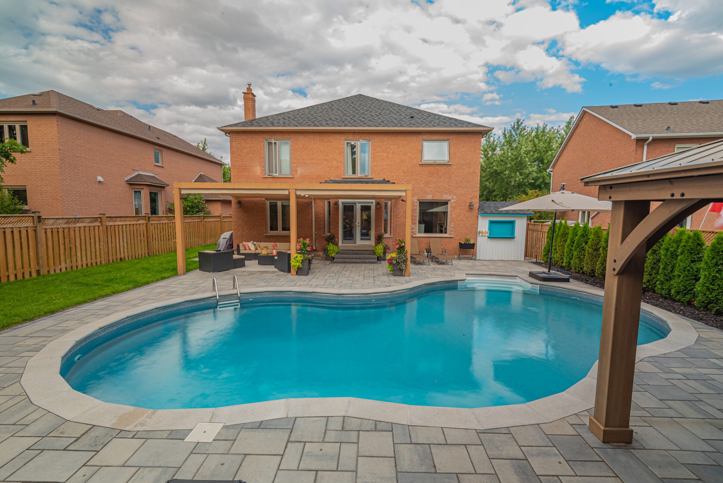 A suburban backyard features a clear pool, wooden pergola with seating, and brick house. Greenery lines the fence, under a partly cloudy sky.