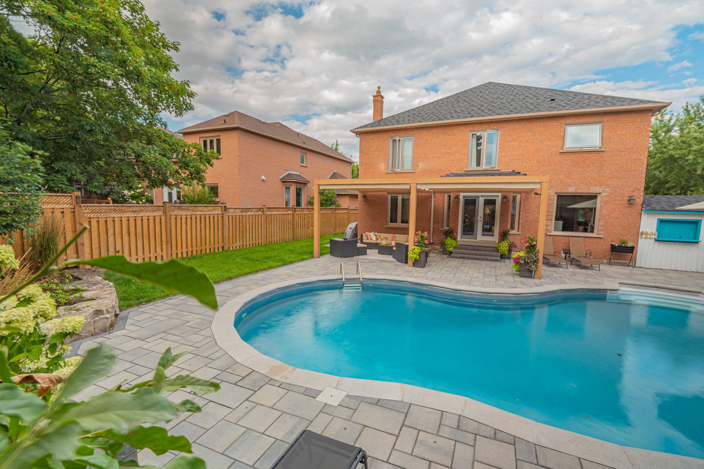 A modern brick house with a wooden pergola, adjacent to a backyard swimming pool, surrounded by greenery and a wooden fence.