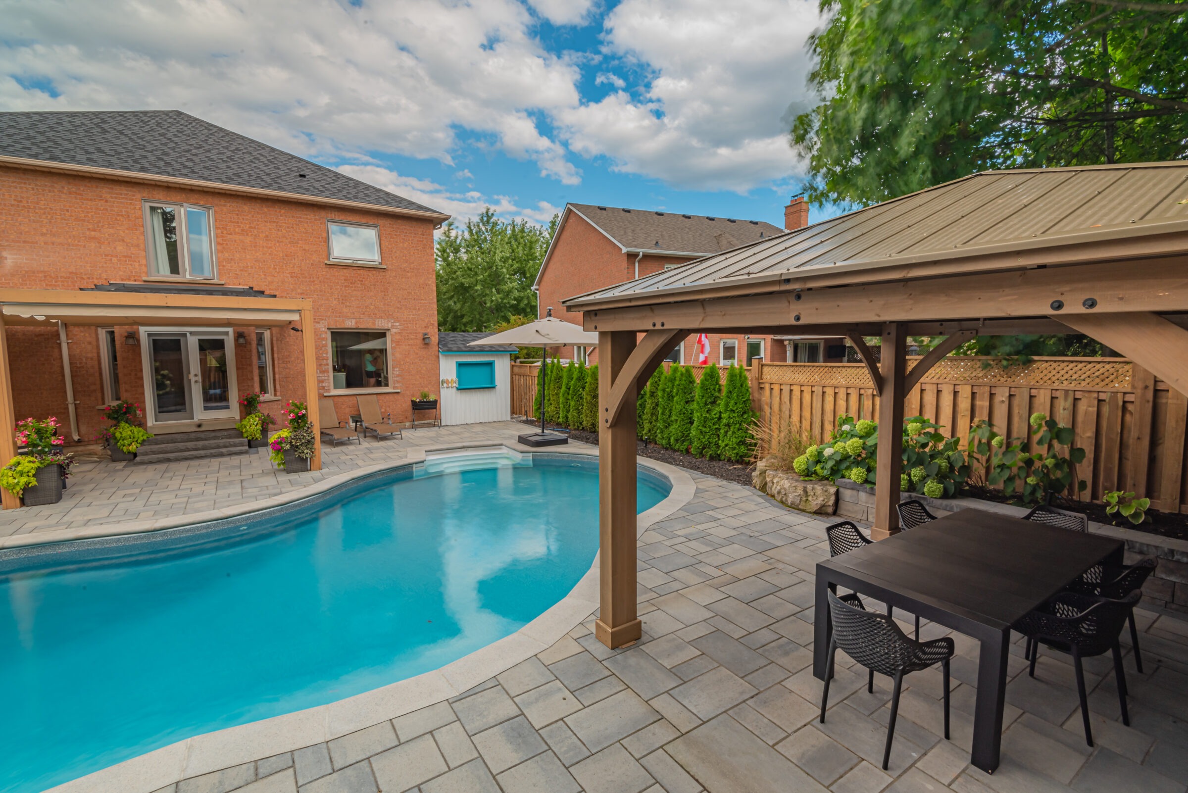 A modern backyard with a brick house, pool, patio, gazebo, and dining area, surrounded by greenery and a wooden fence.