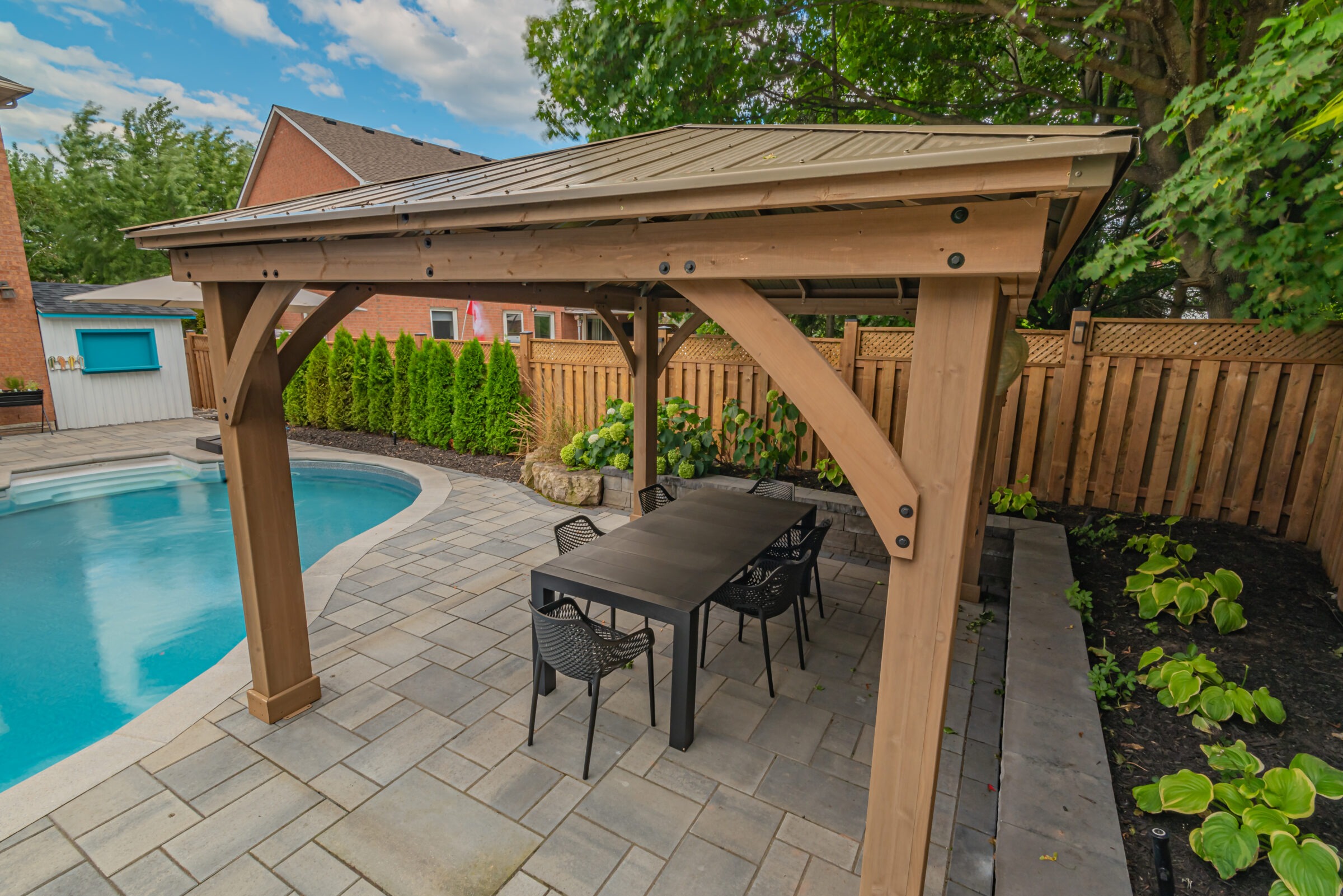 A backyard with a pool, wooden pergola, dining table, chairs, plants, and fenced area, surrounded by brick houses and cloudy sky.