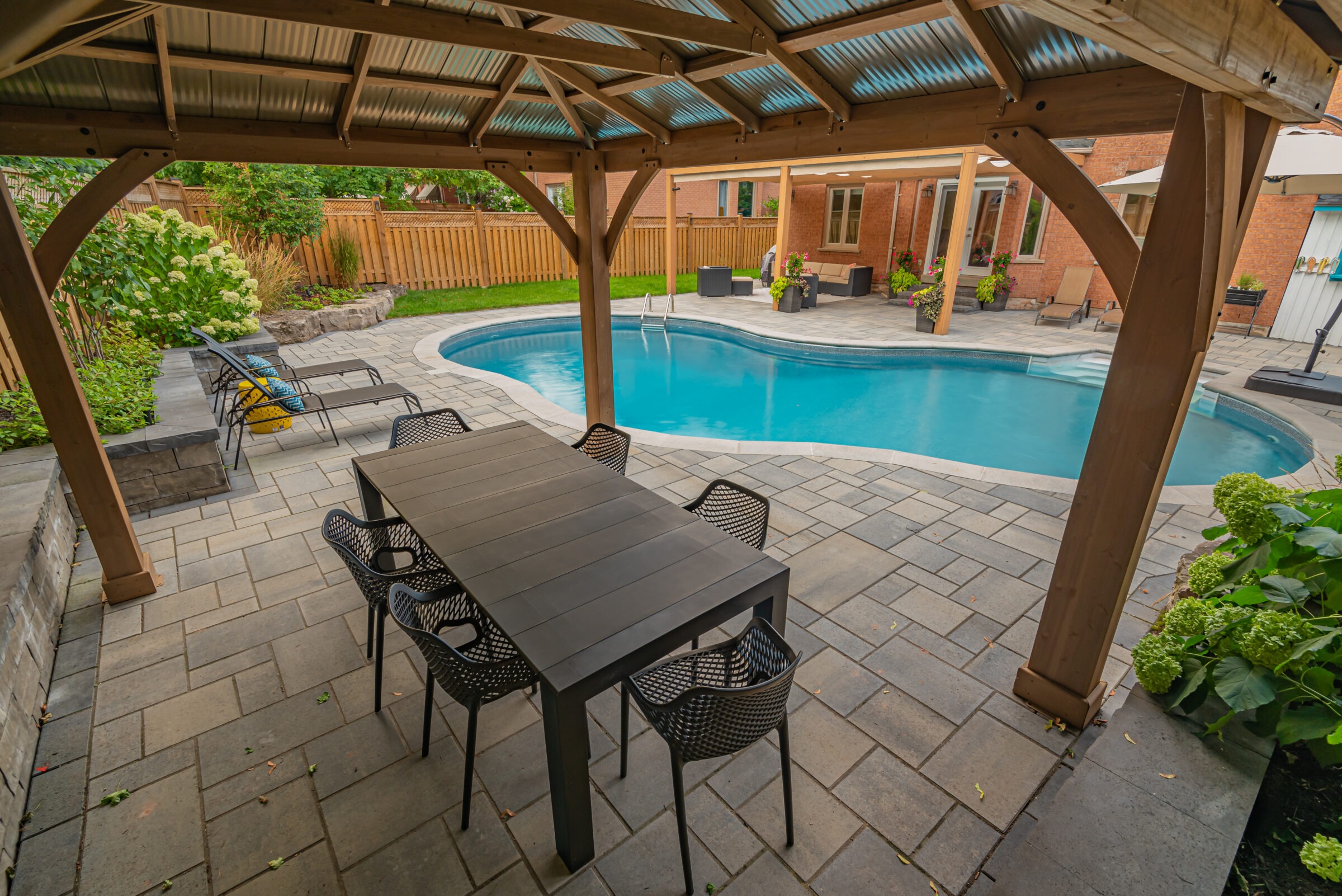 Covered patio with dining table overlooking a backyard pool, lounge chairs, and greenery. Person in the distance near house.