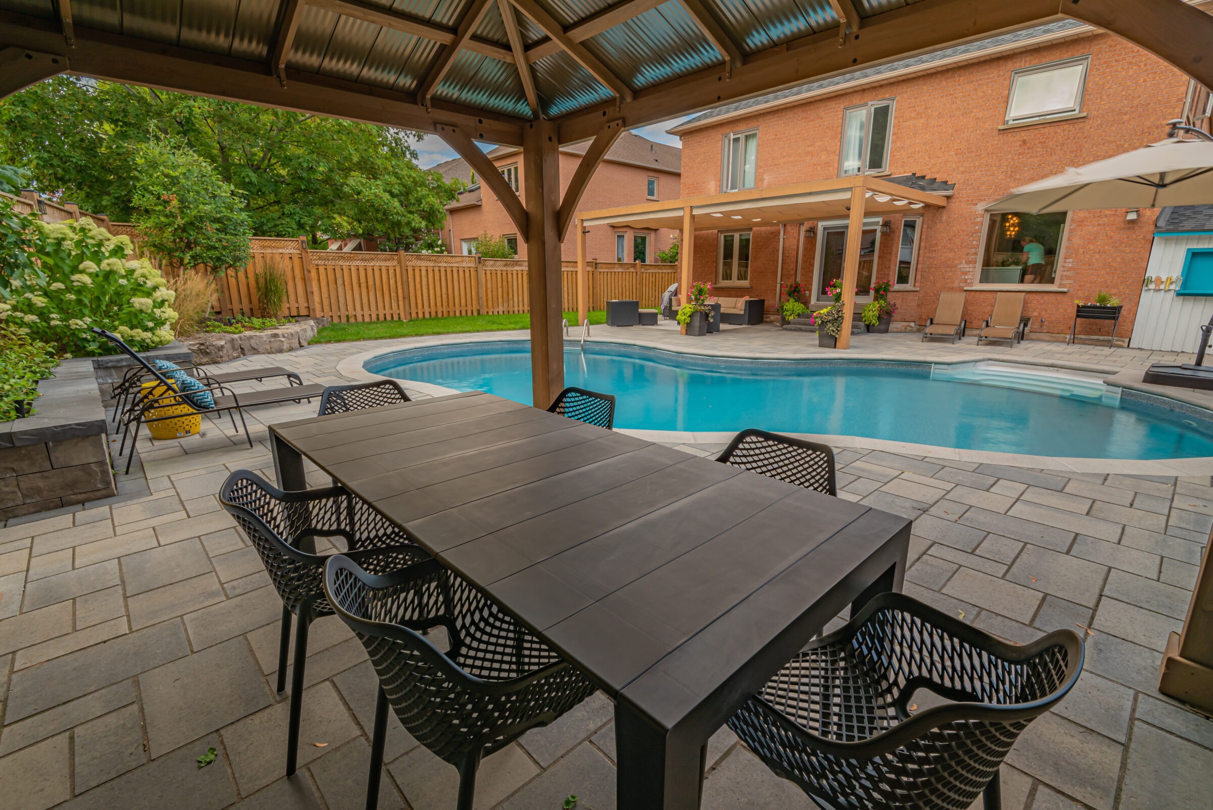 A backyard patio with a dining table, chairs, swimming pool, and a brick house. Lush greenery and lounge chairs are visible.