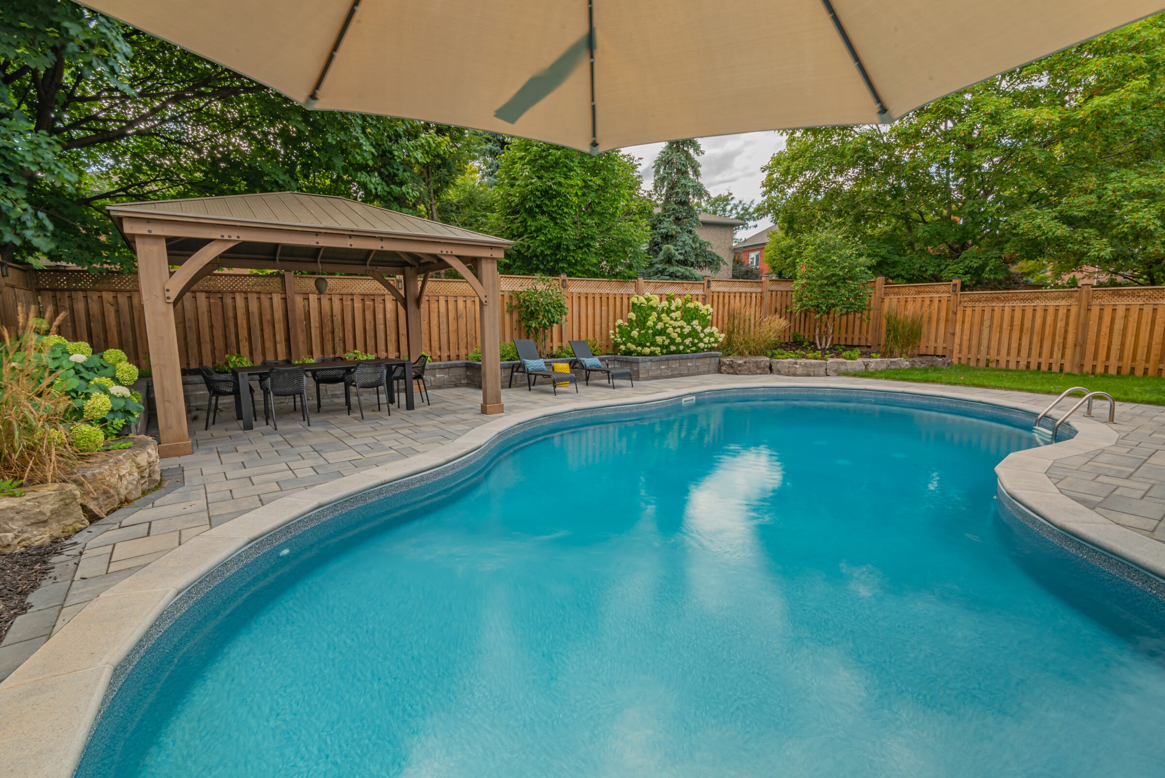 A backyard scene featuring a pristine pool, wooden pergola with seating, surrounded by lush greenery and a wooden fence.