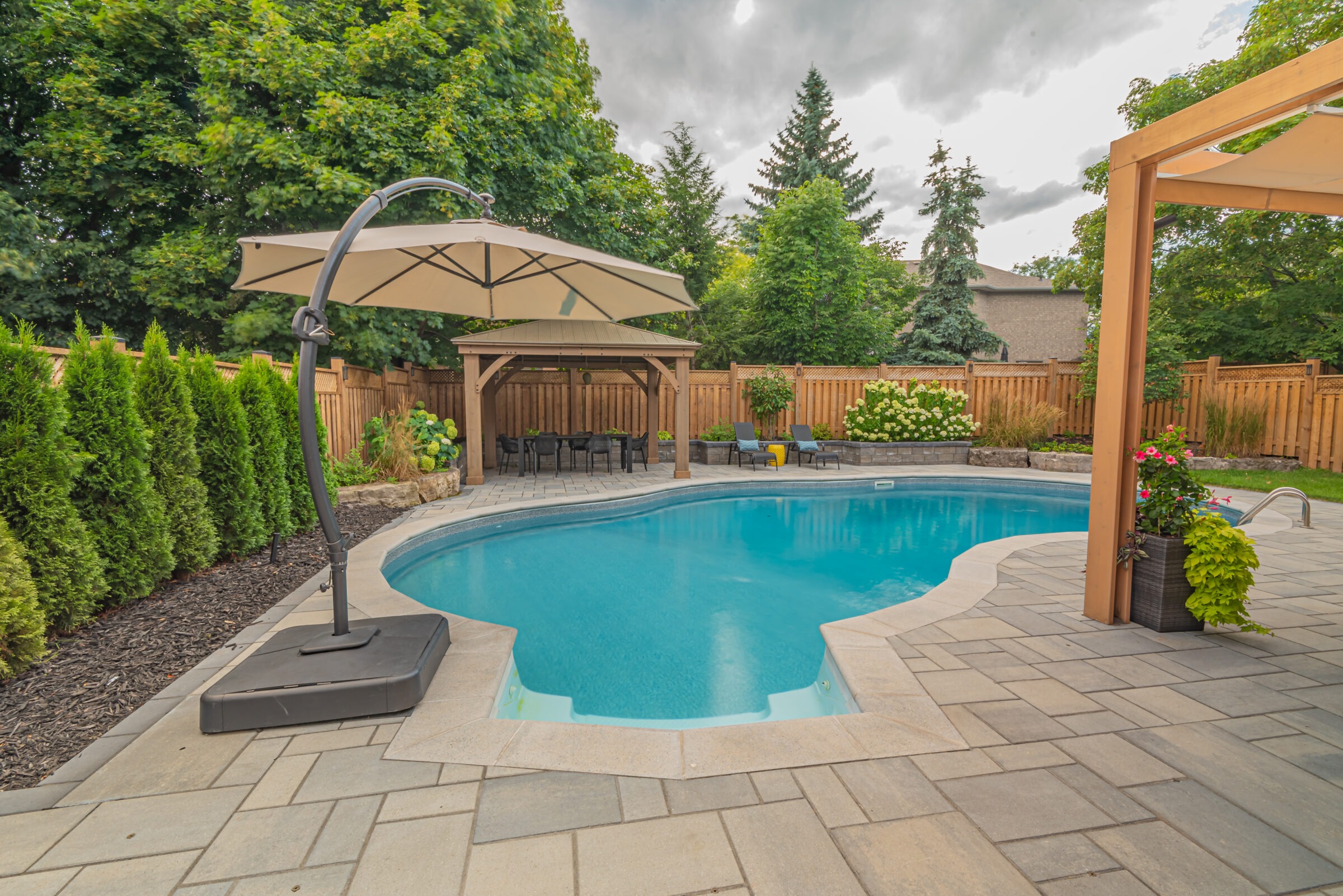A serene backyard features a pool, patio with seating, umbrella, and lush trees, surrounded by a wooden fence with vibrant flowers.