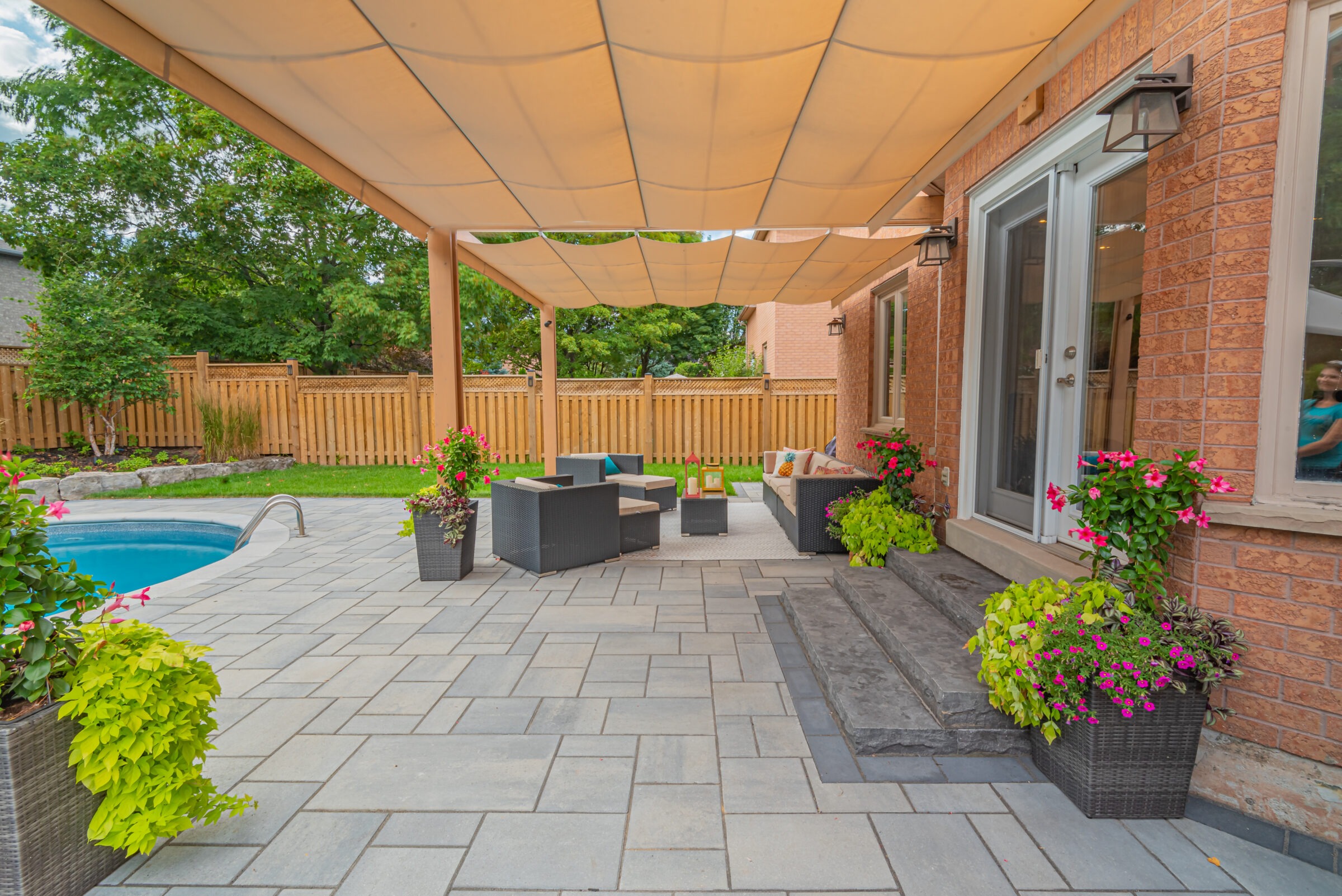 A cozy backyard patio features modern outdoor furniture, vibrant plants, a small pool, and a beige canopy against a wooden fence backdrop.