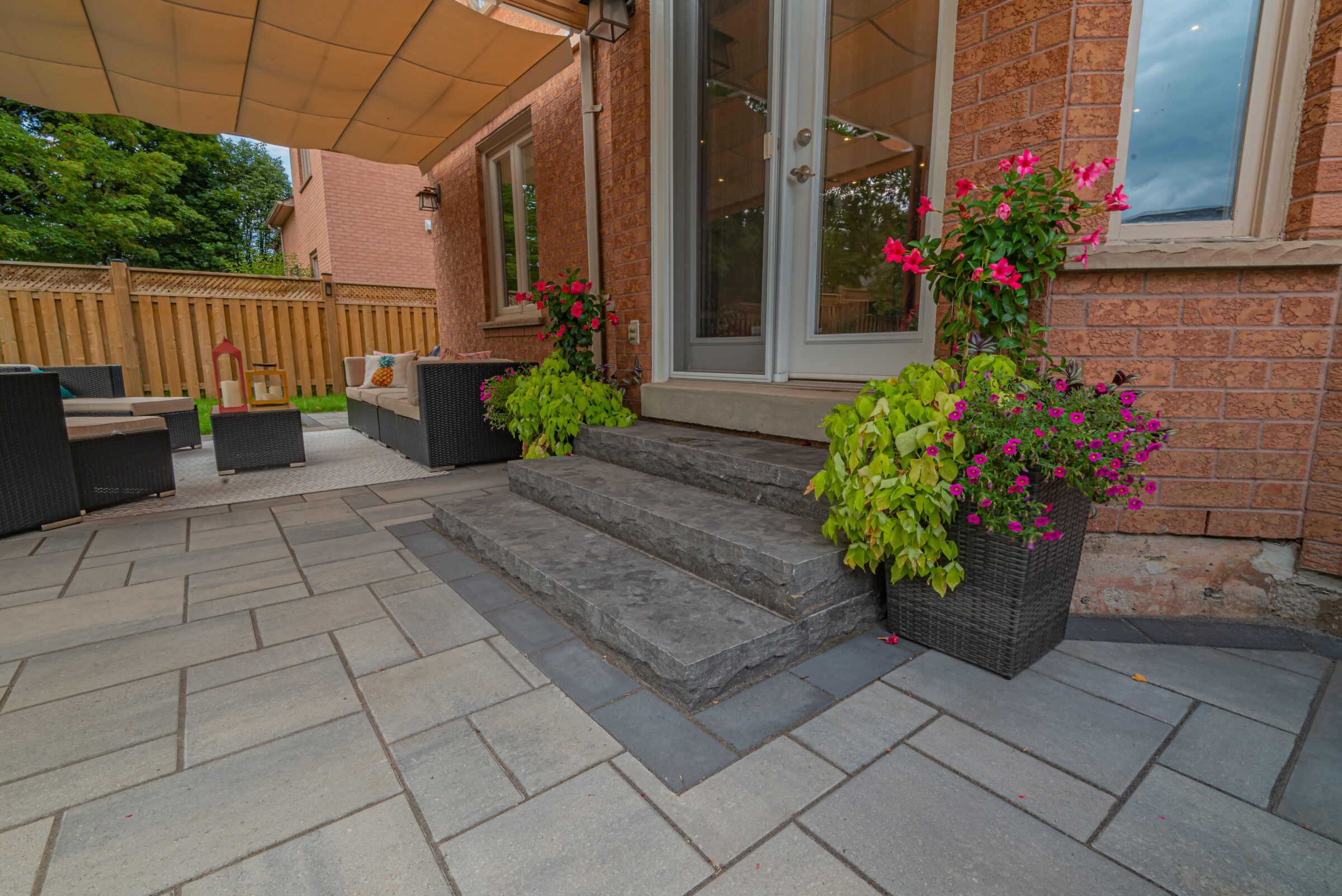 A cozy backyard patio with stone steps, wicker furniture, colorful flower pots, and a wooden fence, creating a peaceful outdoor space.