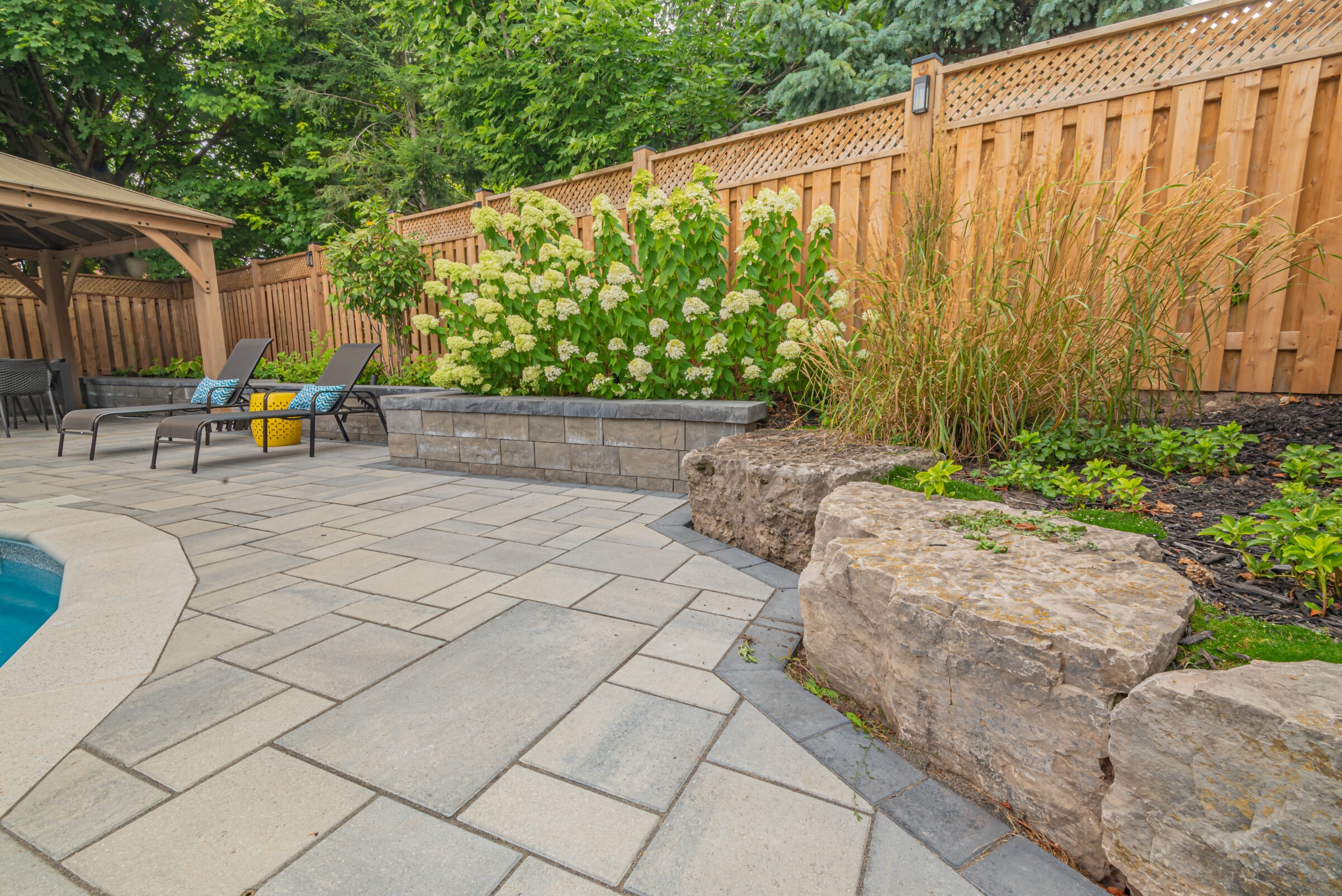 A serene backyard features a stone patio, lush greenery, a wooden fence, and a cozy seating area beside a pool.