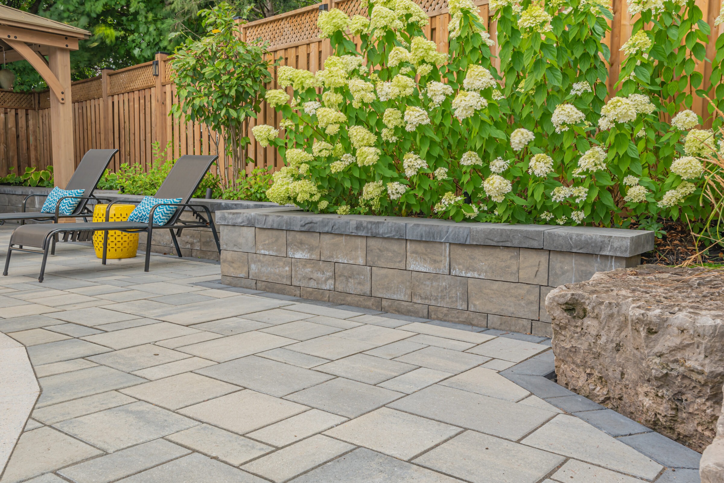 A peaceful backyard patio with two lounge chairs, bright cushions, lush green plants, stone tiles, wooden fencing, and a serene ambiance.