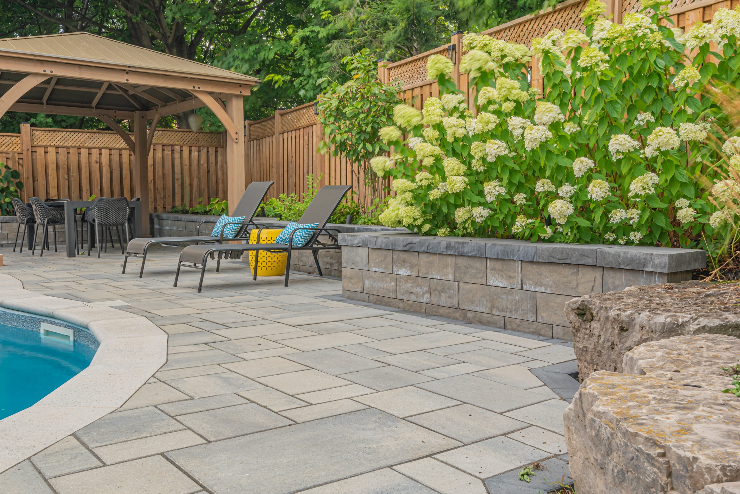 A backyard patio features a pool, lounge chairs, a gazebo, and lush greenery against a wooden fence, creating a relaxing outdoor space.