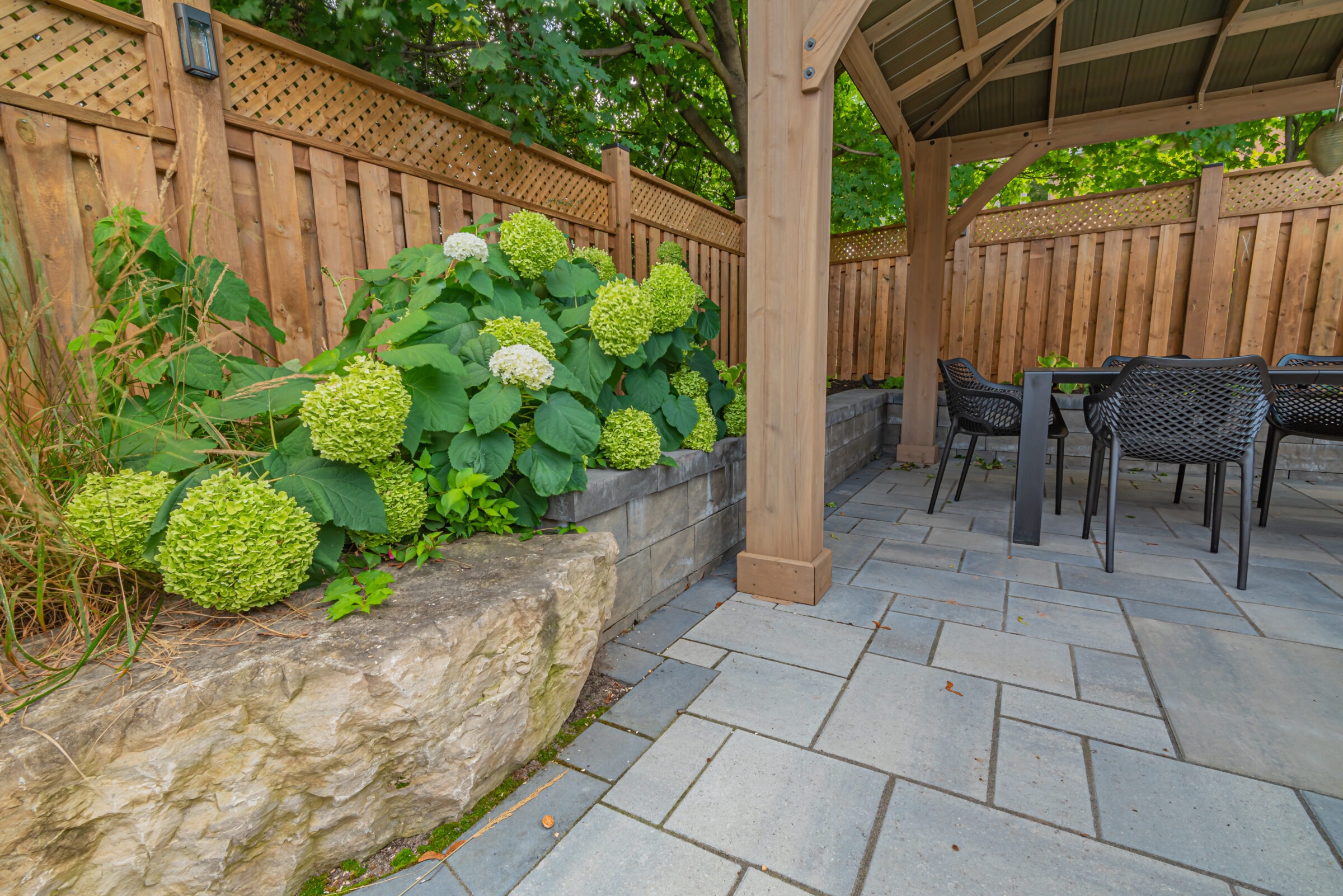 A cozy patio with hydrangeas, wooden pergola, stone tiles, surrounded by a wooden fence. Black chairs around a table complete the seating area.