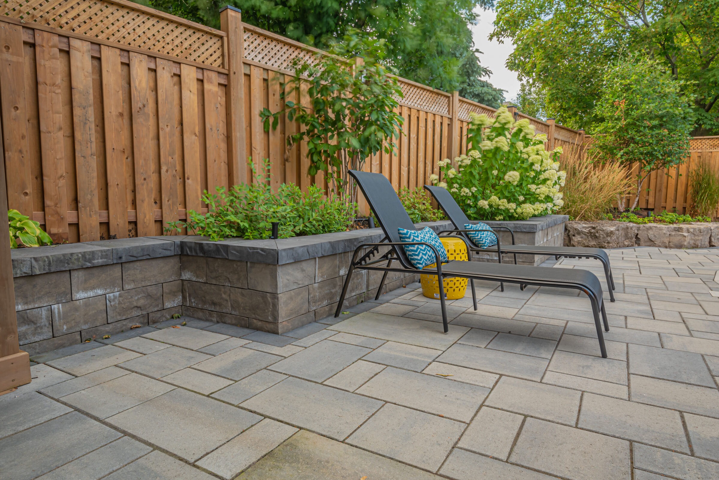 Two lounge chairs with colorful cushions on a paved patio, surrounded by lush greenery and wooden fencing, creating a serene garden space.
