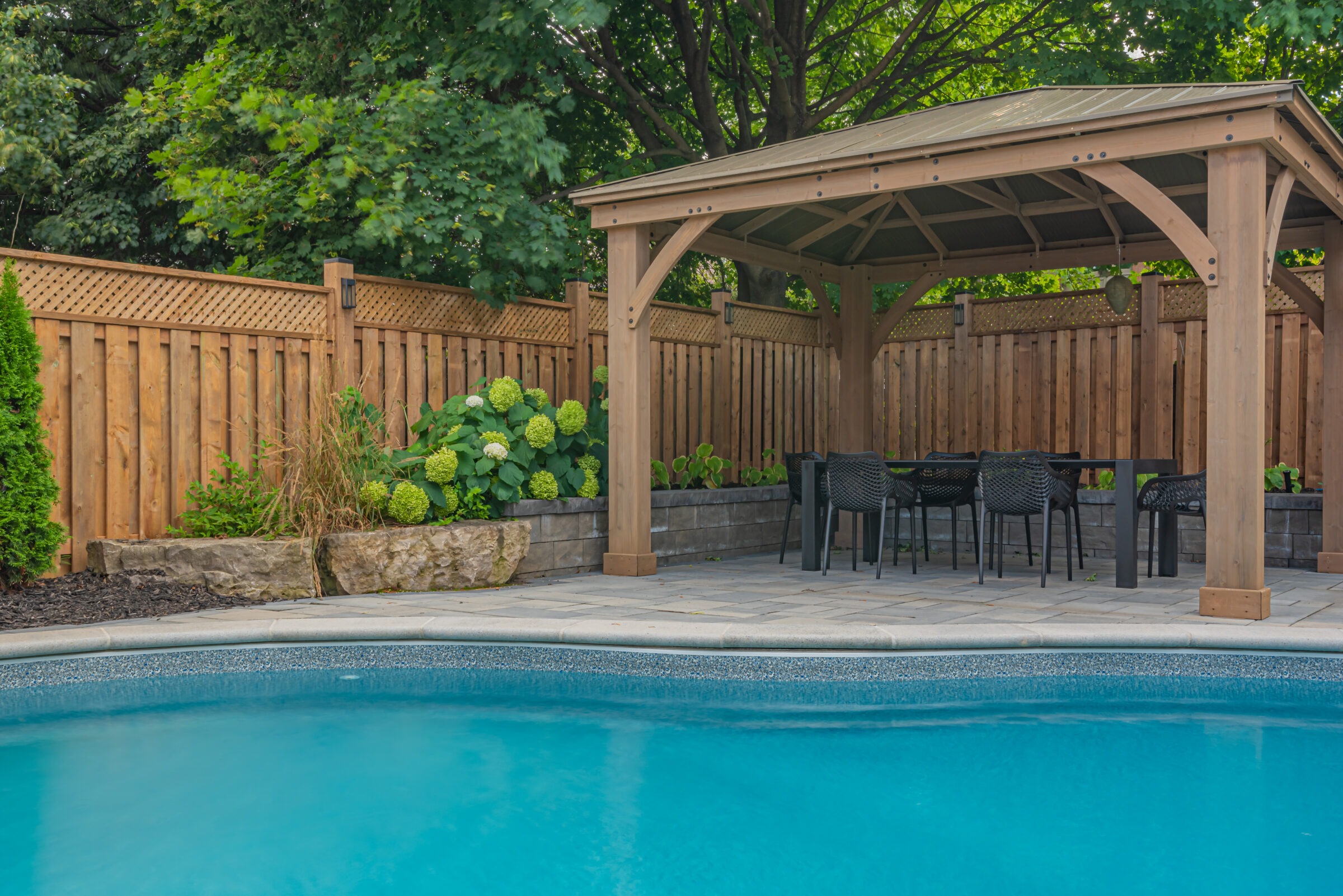A backyard pool with clear water is bordered by a wooden pergola, surrounded by lush greenery and a wooden fence.