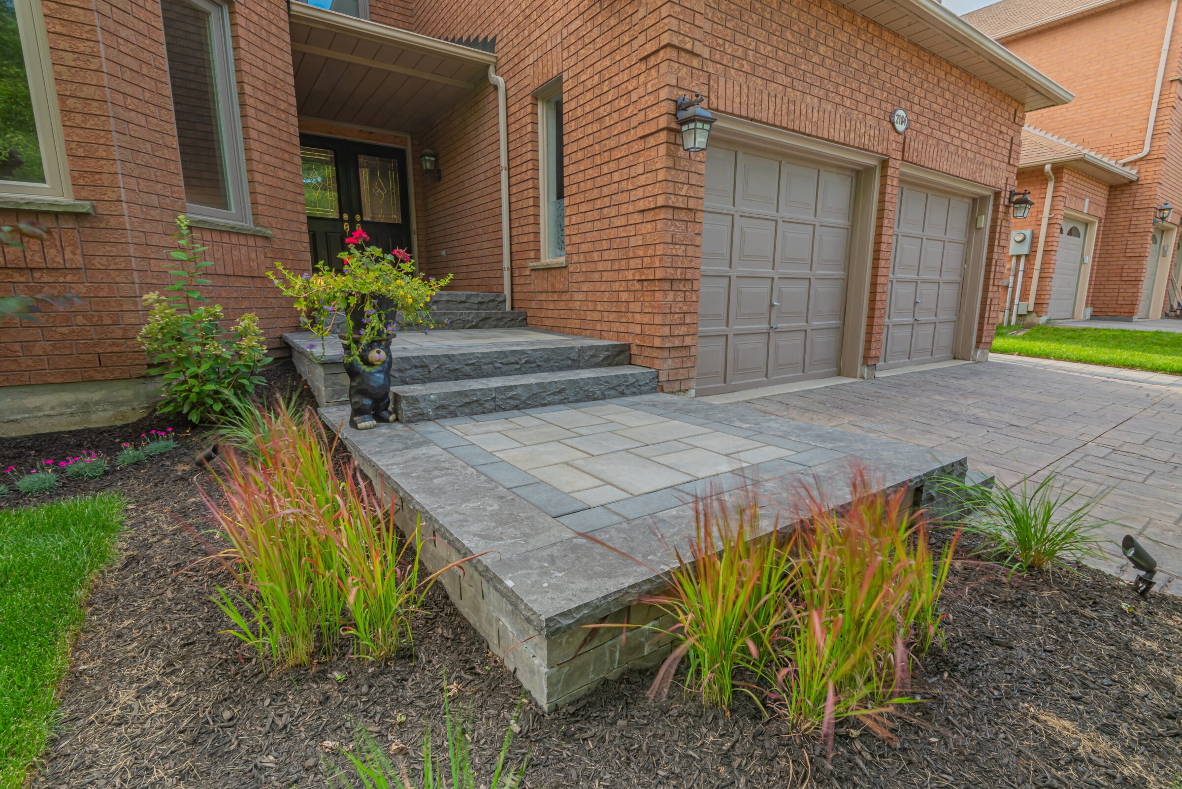 Red brick house with a two-car garage, landscaped entrance, and small bear statue. Well-maintained garden with shrubs and grass.