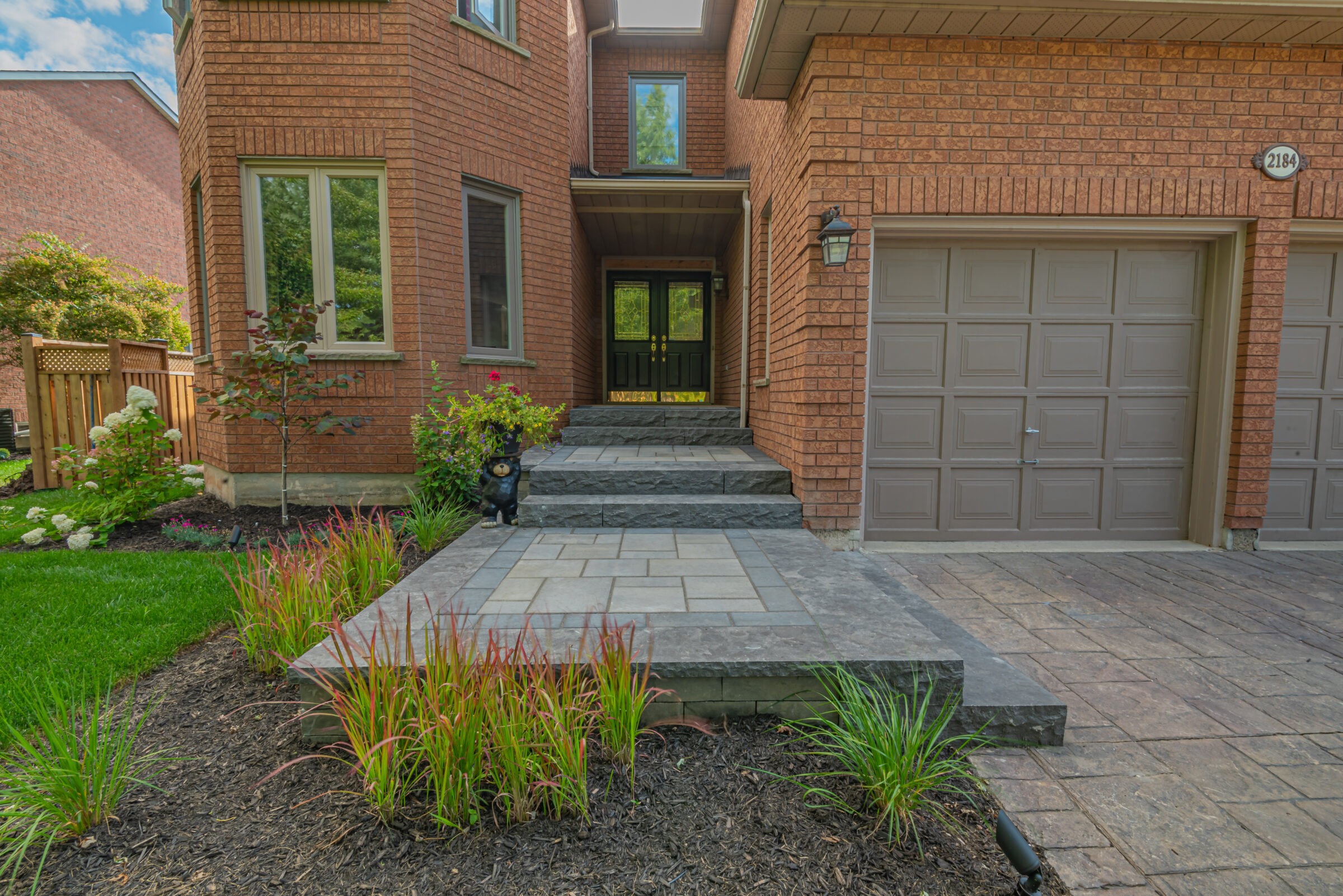 A brick house with a two-car garage, front steps, and a well-maintained garden featuring decorative plants and a small bear sculpture.
