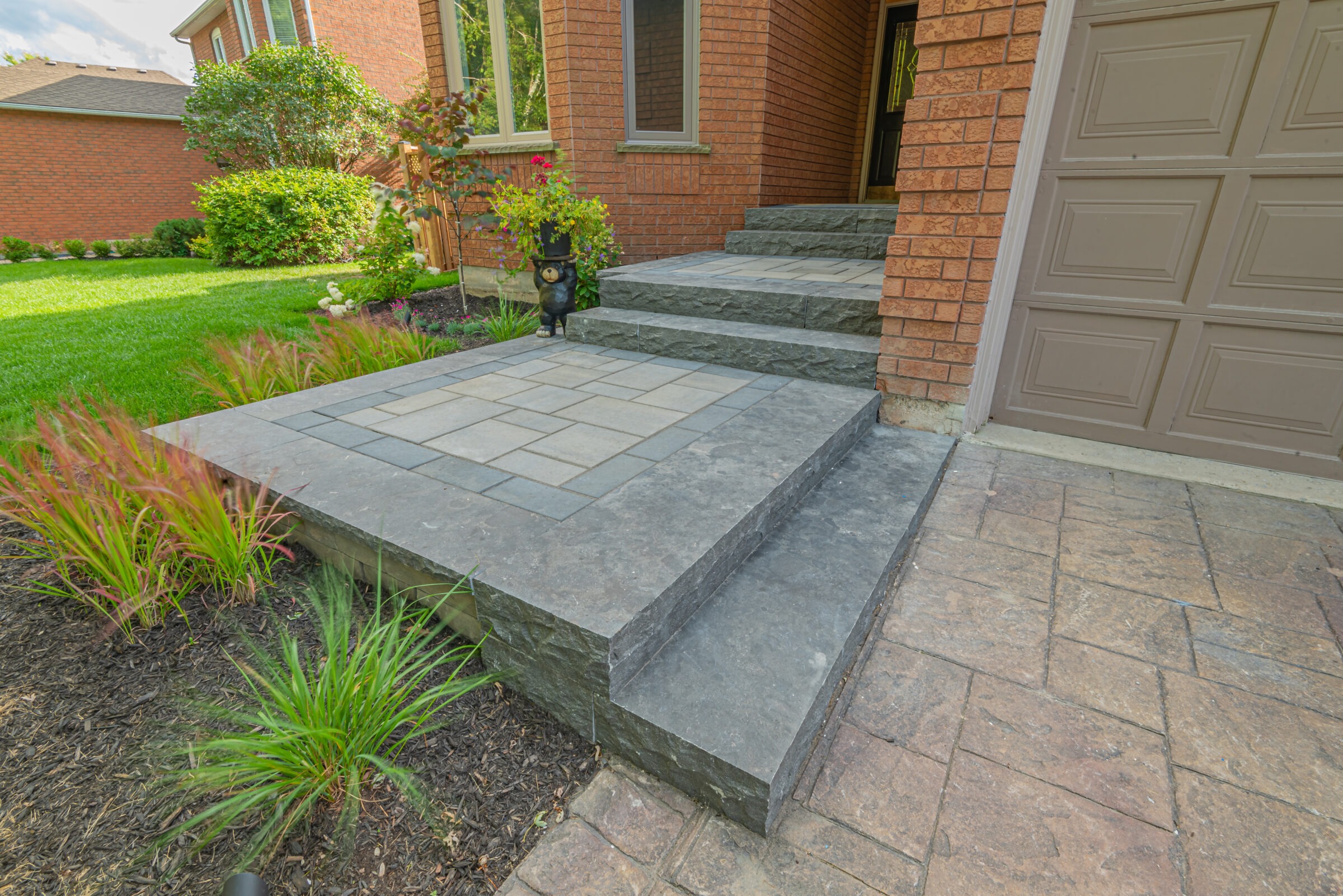 Brick house entrance with stone steps, landscaped garden, grass, and a small bear statue wearing a top hat near the front door.