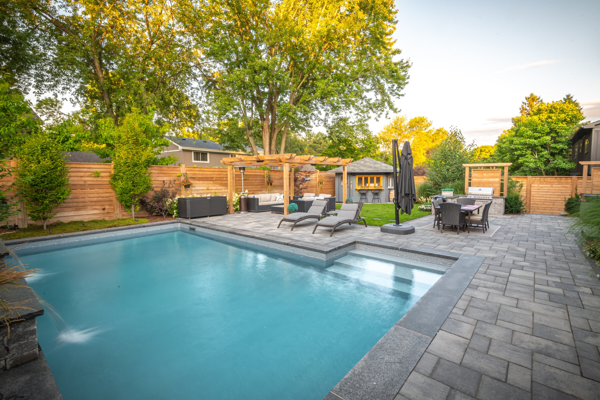A serene backyard with a swimming pool, lounge chairs, pergola, outdoor dining area, and lush greenery, surrounded by a wooden fence.