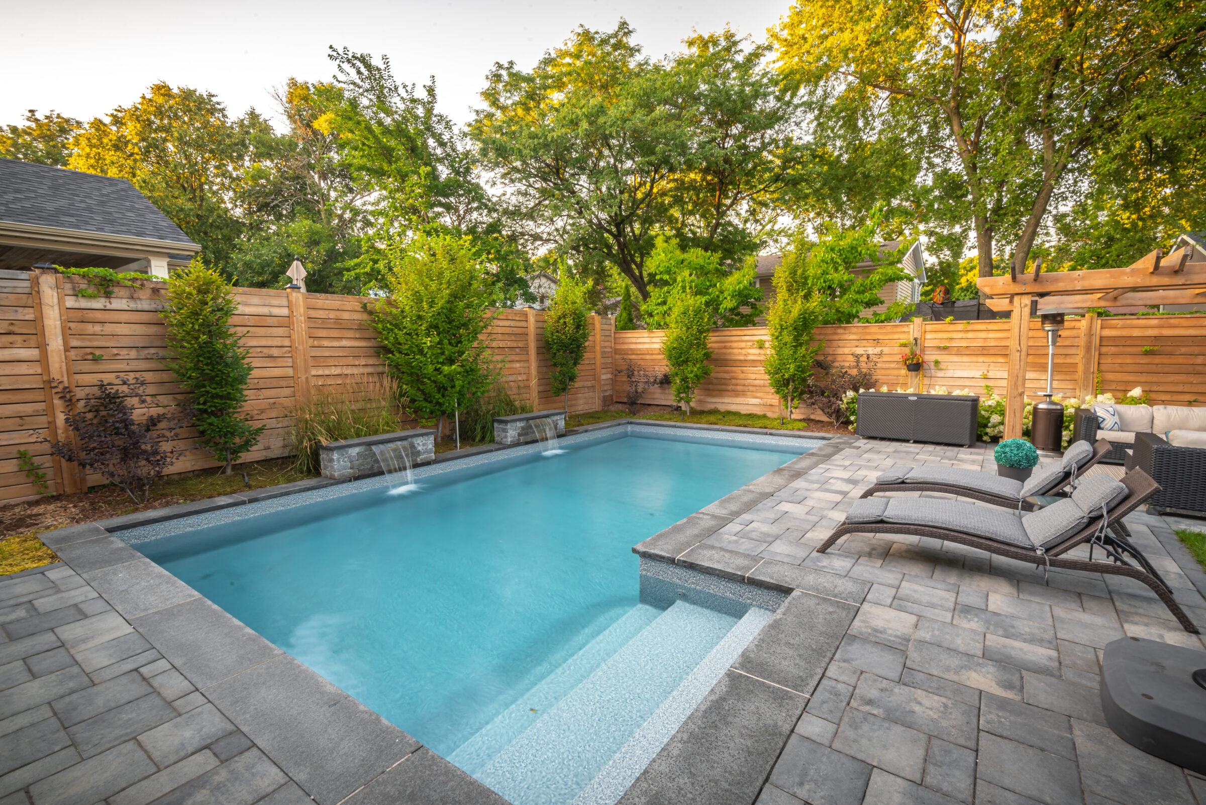 A serene backyard features a rectangular pool, lounge chairs, and wooden fencing, surrounded by lush green trees and well-manicured plants.