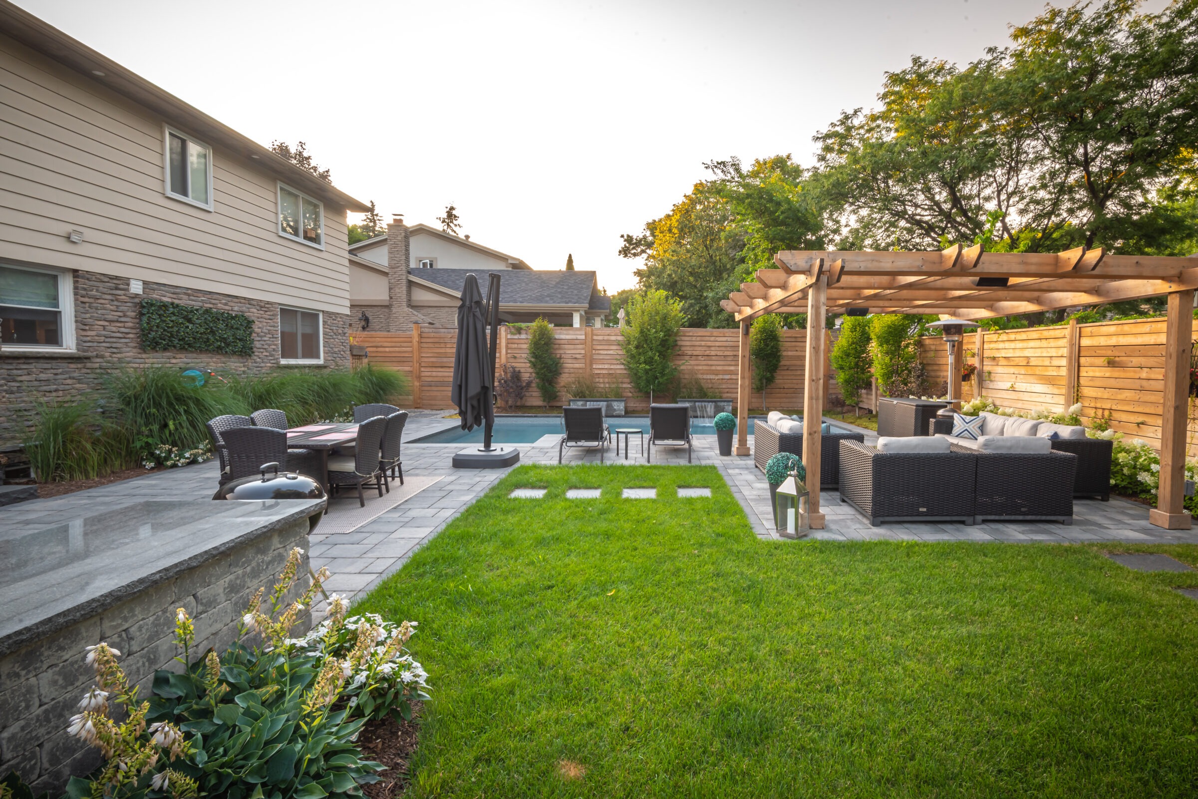 A modern backyard features a pergola, outdoor seating, dining area, and lush greenery, creating a relaxing and inviting atmosphere for gatherings.