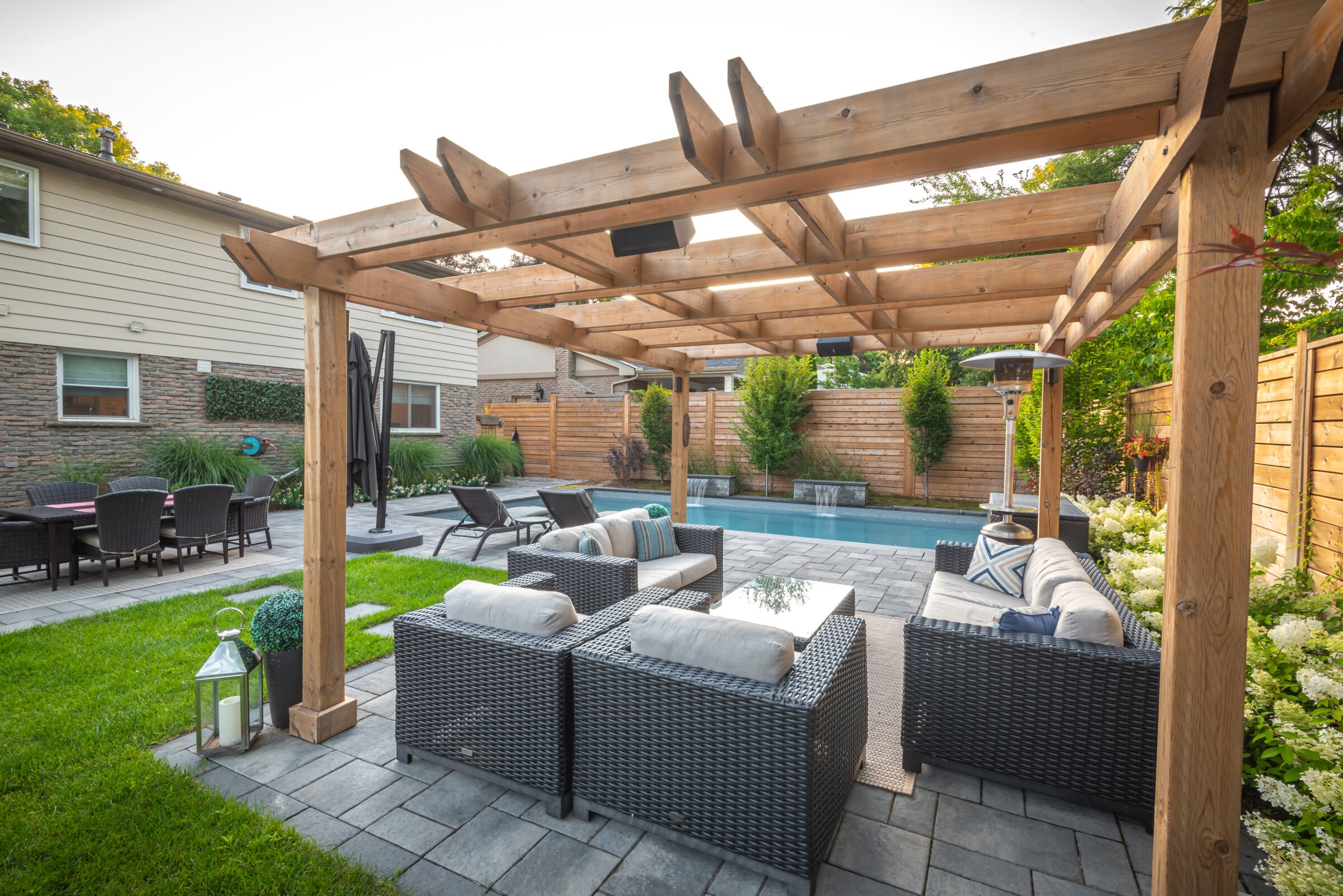 A backyard features a wooden pergola, wicker patio furniture, a small pool, neatly manicured lawn, and surrounding wooden privacy fence.