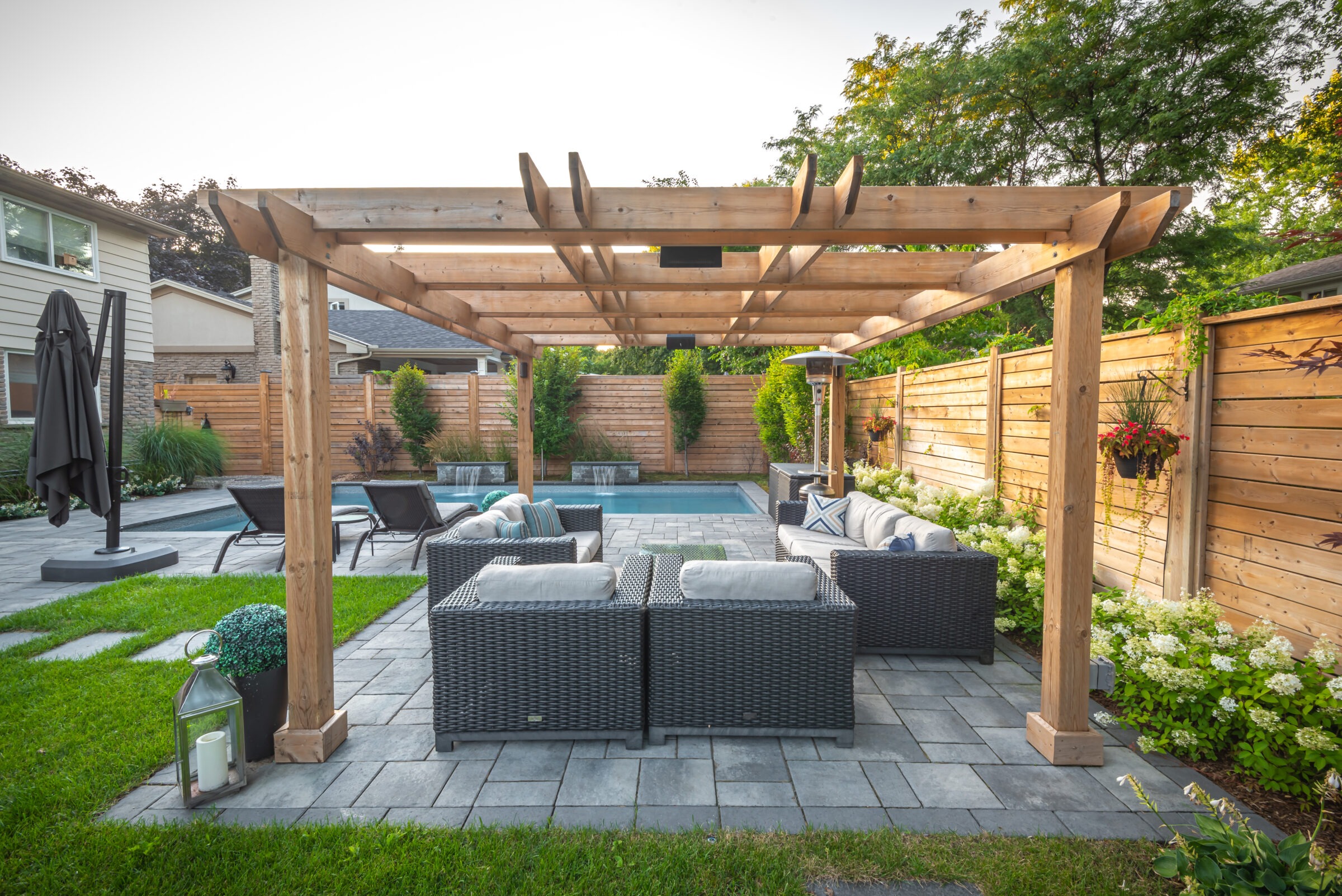 A backyard patio features a wooden pergola, comfortable seating, potted plants, and a pool surrounded by a neatly organized garden and wooden fence.