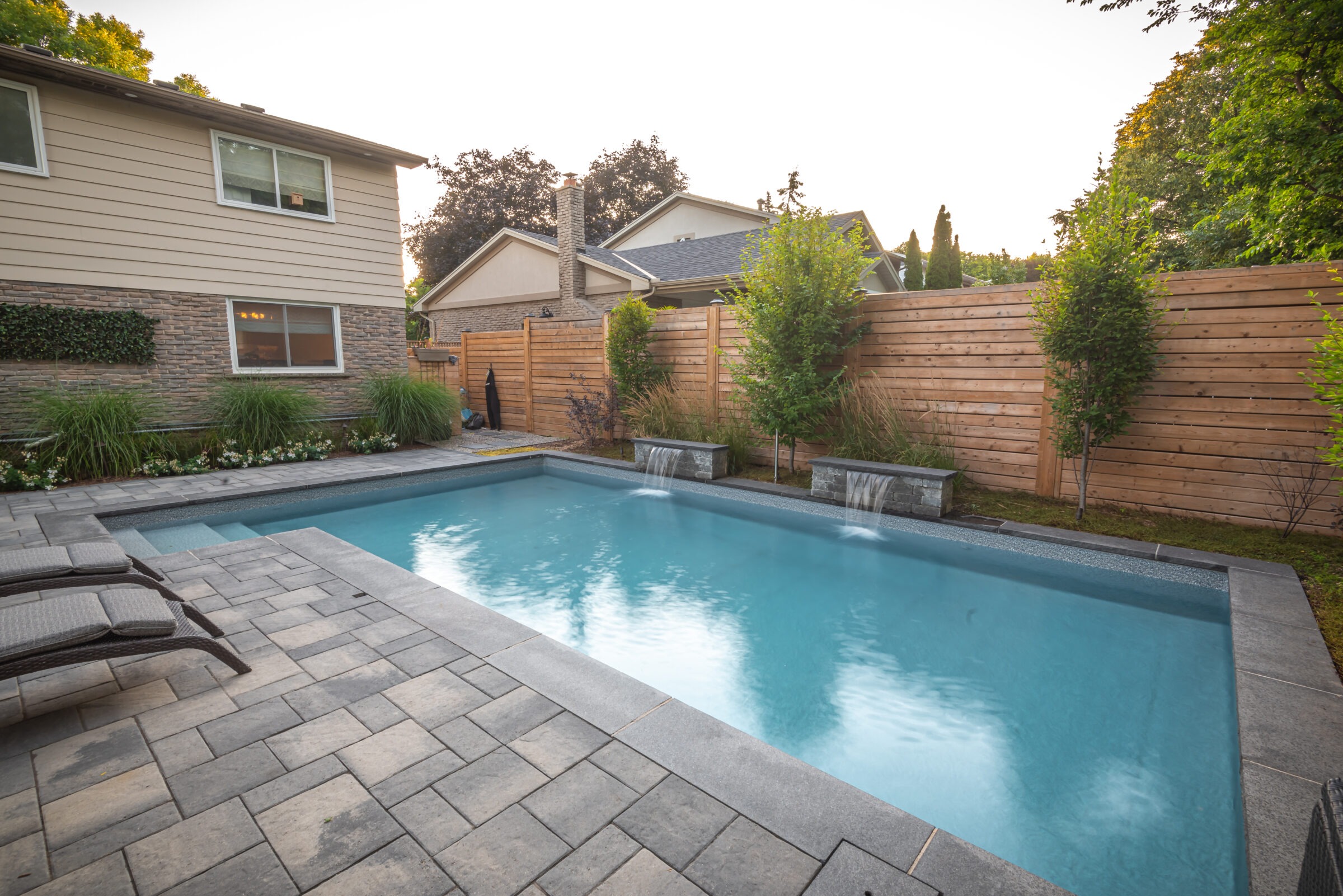 Modern backyard with a rectangular swimming pool, stone patio, wooden fence, and lush greenery, adjacent to a residential house at sunset.