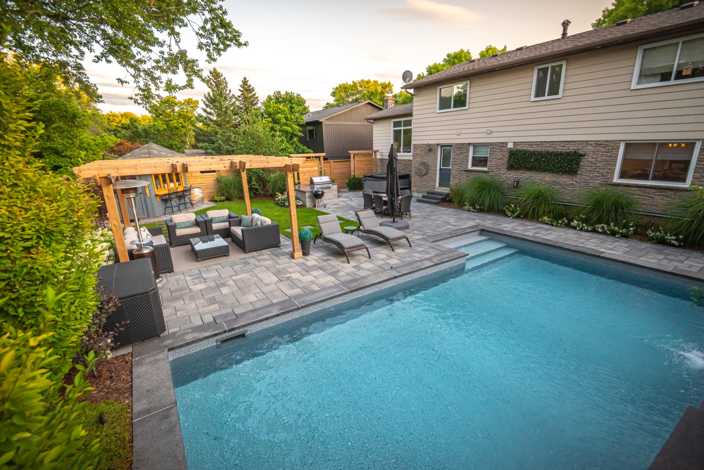 Backyard with a pool, pergola, and seating area. A house is visible, surrounded by trees and landscaped greenery. Peaceful outdoor setting.