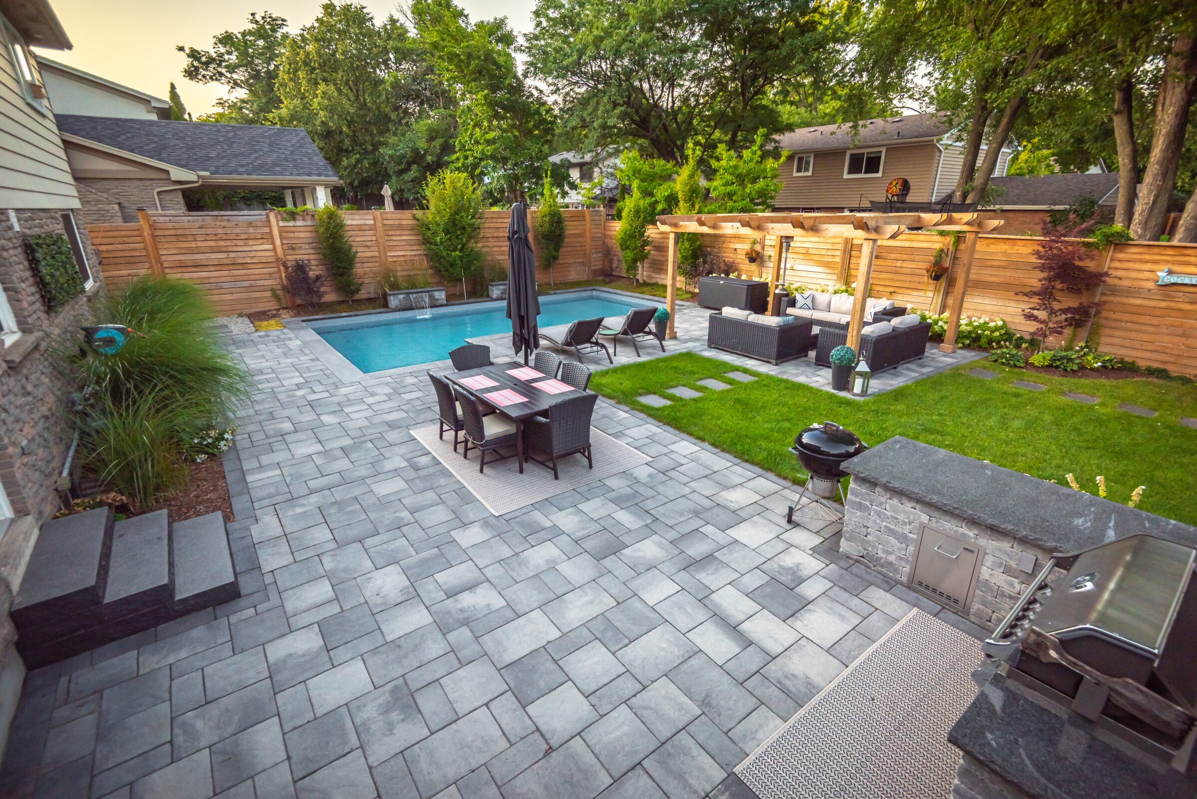 Modern backyard features a pool, outdoor dining, and lounge area under a pergola, surrounded by greenery and stone pavement.