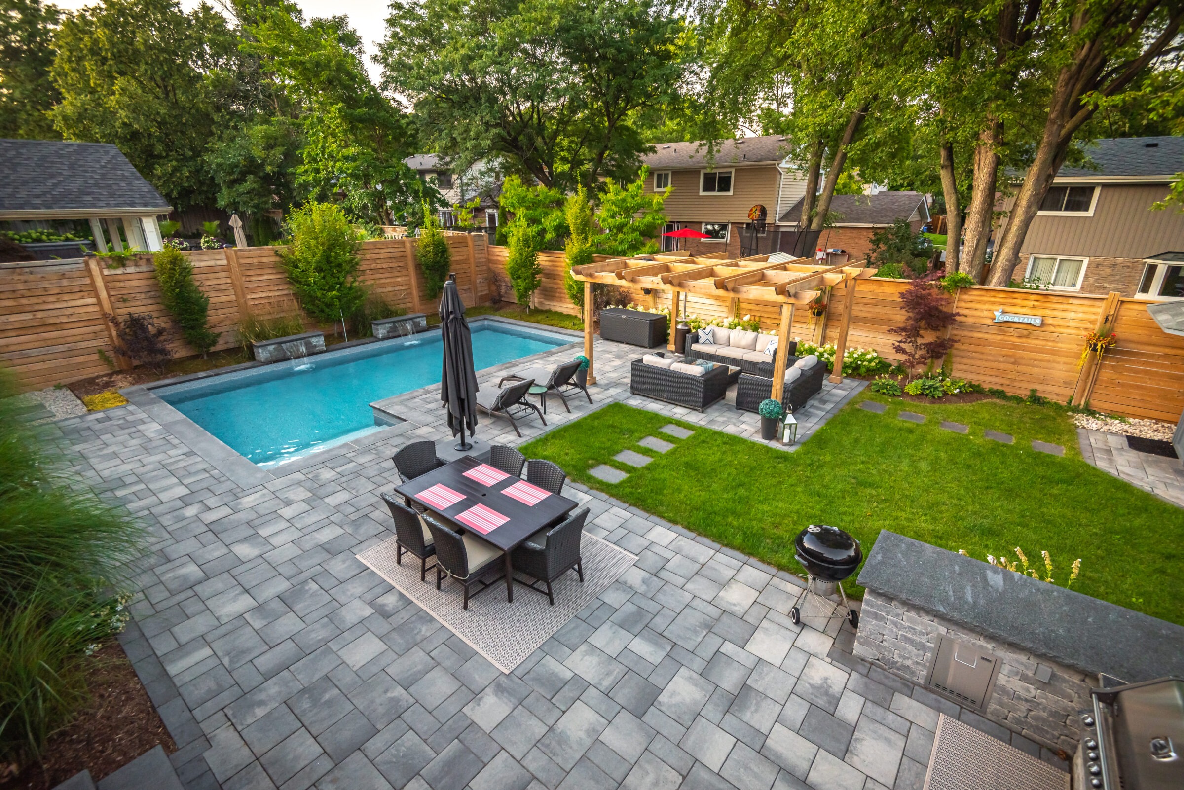 A spacious backyard features a swimming pool, patio furniture, pergola, grill, and lush greenery, surrounded by wooden fences and nearby residential houses.