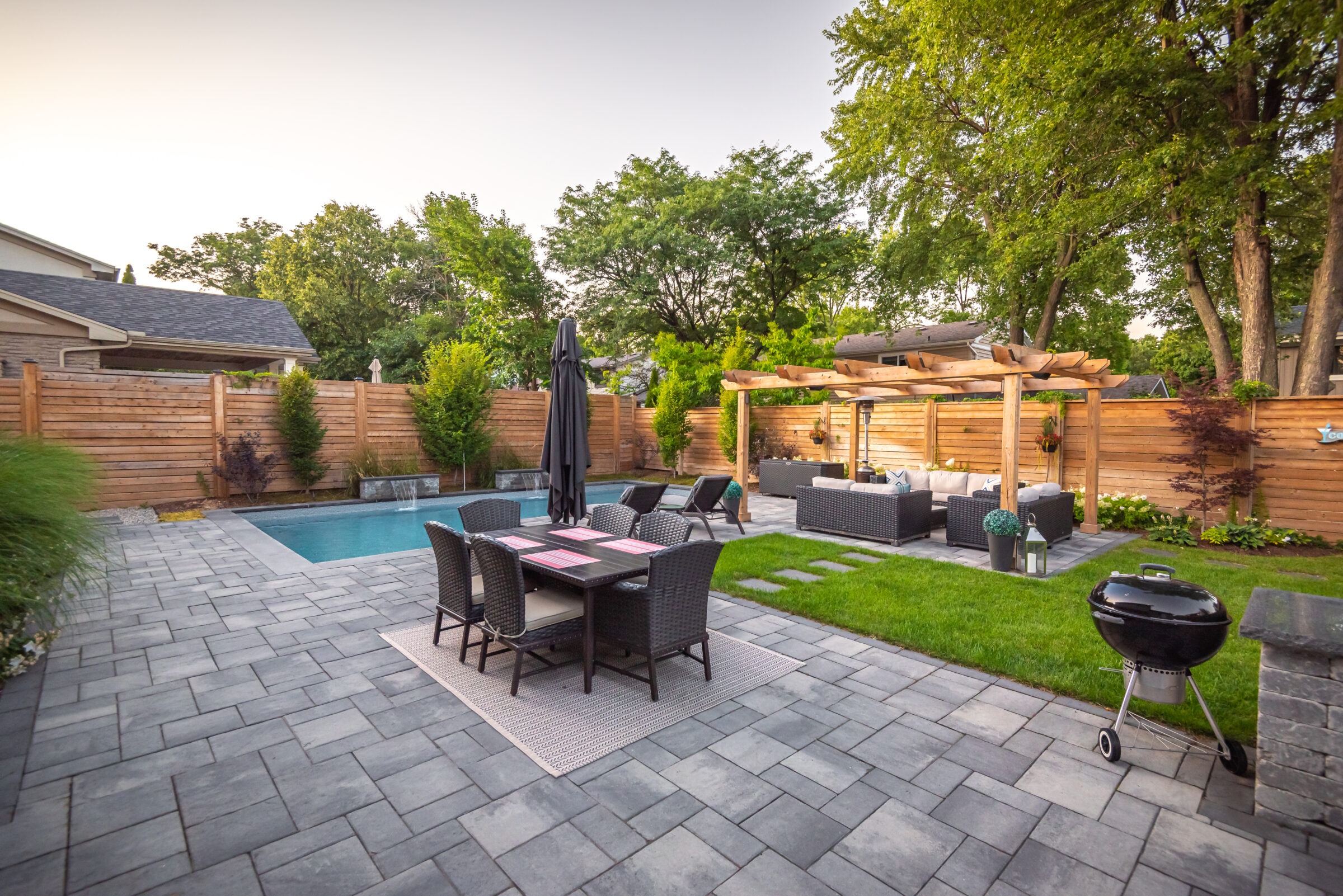 Modern backyard featuring a pool, patio dining set, pergola with seating, and greenery, enclosed with a wooden fence under clear skies.