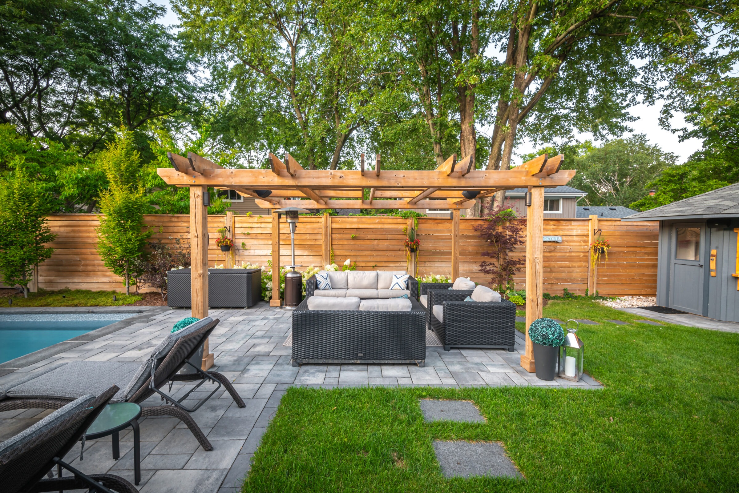 A backyard oasis with a wooden pergola, wicker furniture, and a pool surrounded by lush greenery and a wooden fence.