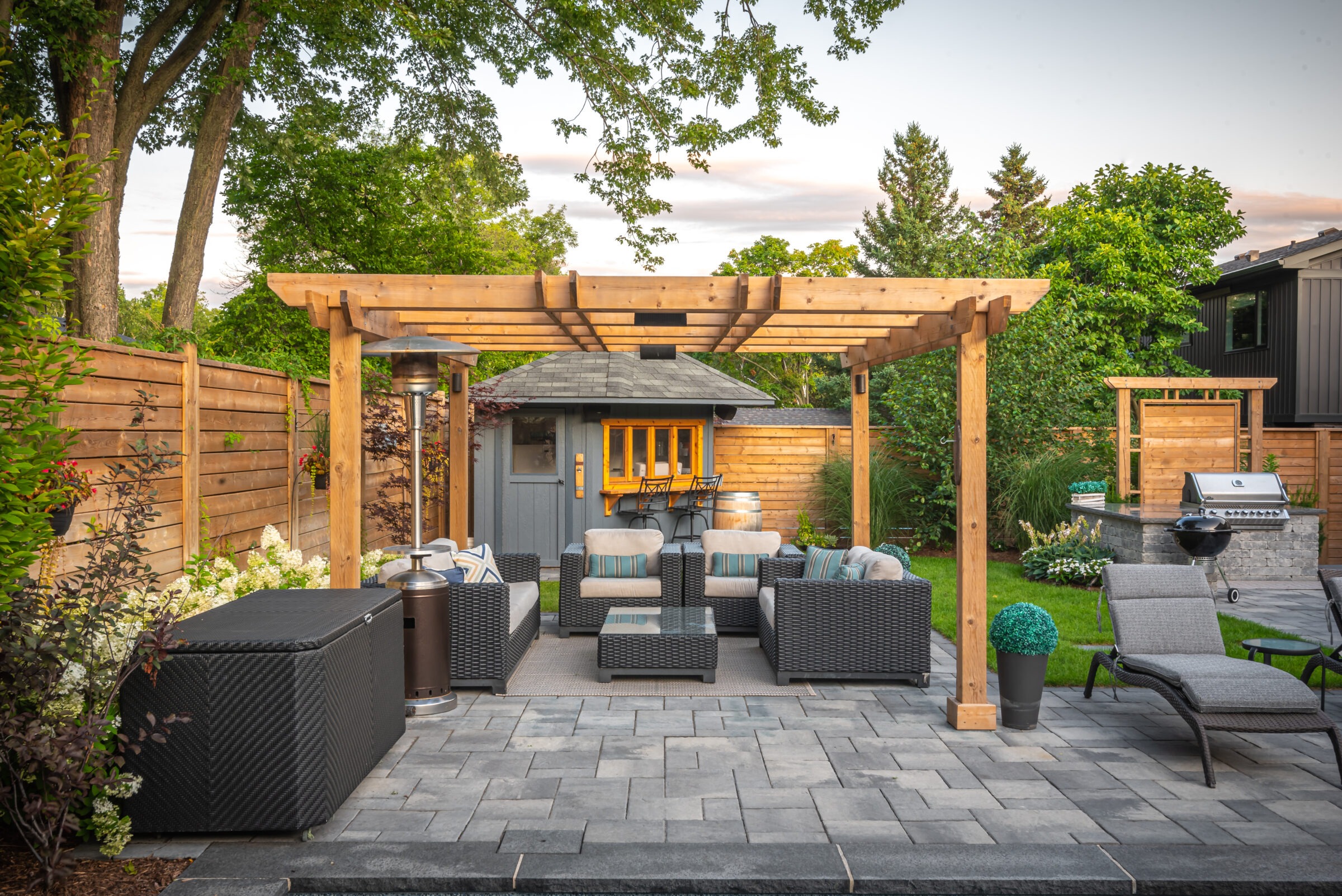 A cozy backyard patio with a pergola, comfortable seating, barbecue grill, and lush green garden surrounded by wooden fencing and a small shed.