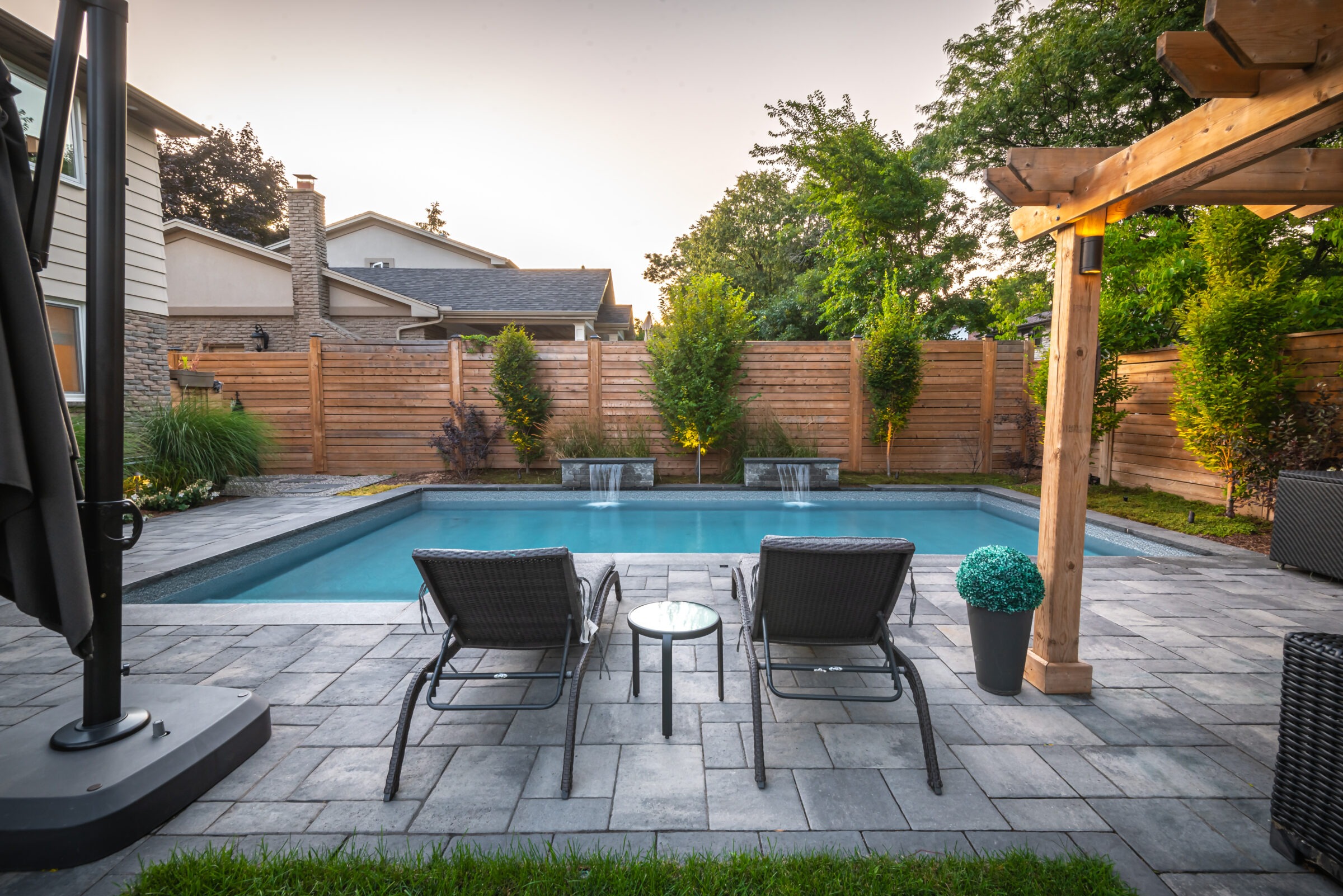 A backyard oasis featuring a serene swimming pool with water features, surrounded by a wooden fence, greenery, and relaxing lounge chairs.