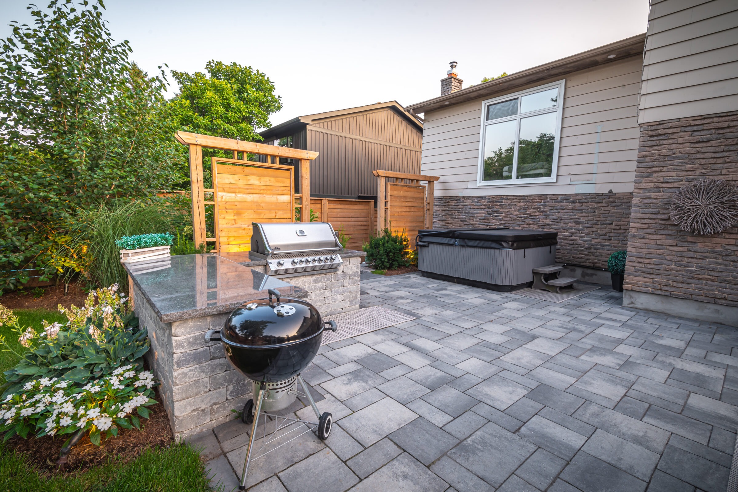 Modern backyard with a barbecue grill, hot tub, and stone patio. Wooden fencing and lush greenery provide a cozy, inviting outdoor setting.