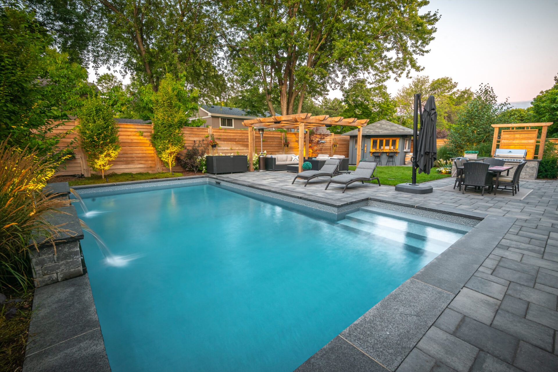 Spacious backyard with swimming pool, lounge chairs, pergola, and dining area. Surrounded by lush greenery and wooden fence, creating a serene retreat.