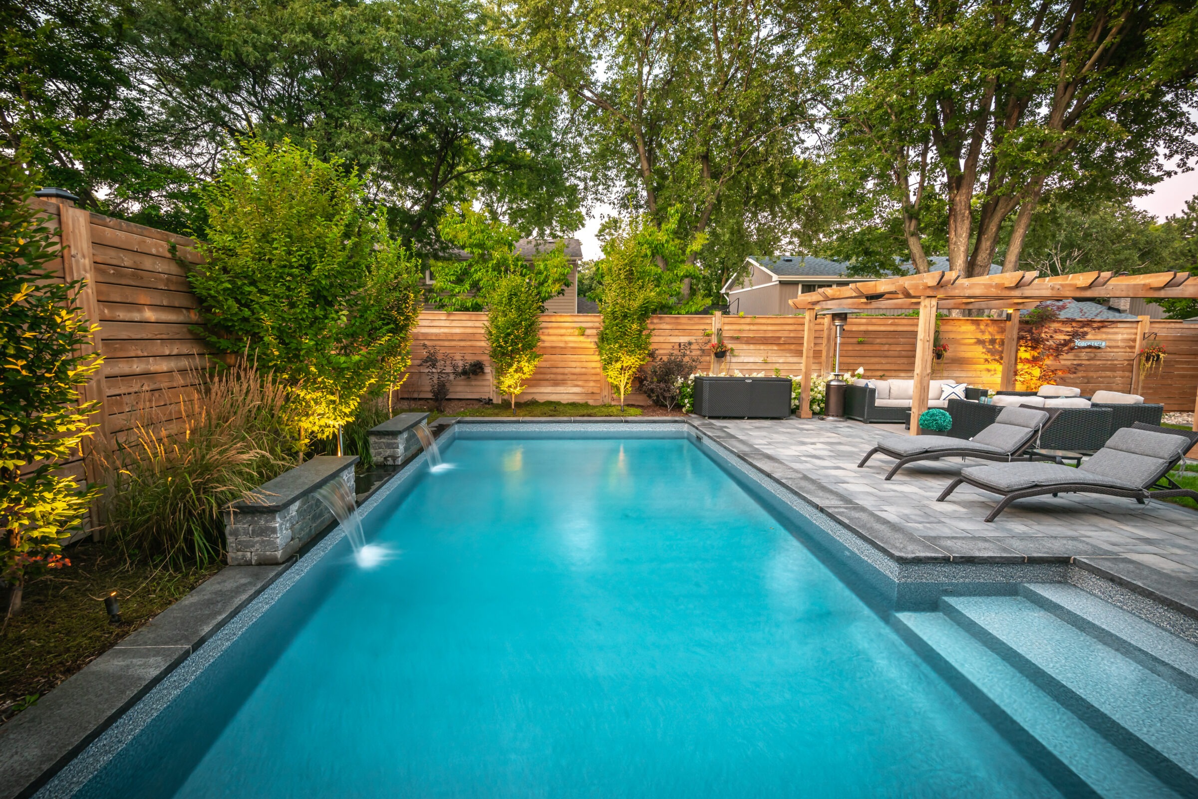 Modern backyard with a rectangular pool, lounge chairs, pergola, wooden fence, and lush greenery, creating a serene and inviting outdoor oasis.