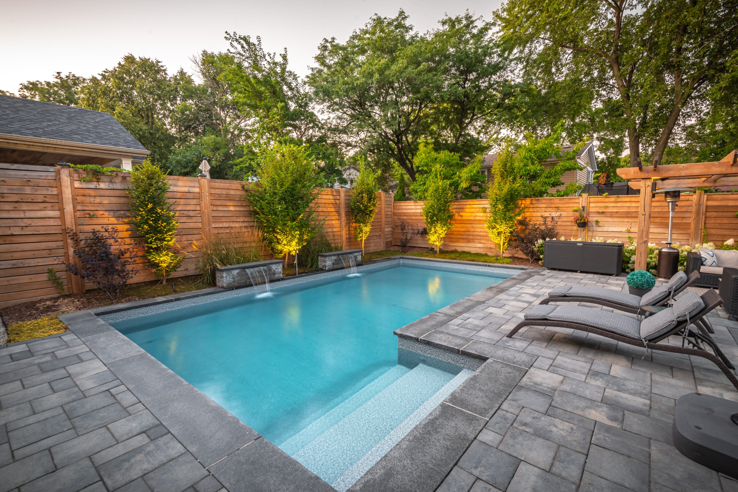 A serene backyard with a modern swimming pool, surrounded by a wooden fence, lush trees, lounge chairs, and ambient lighting highlighting the landscape.