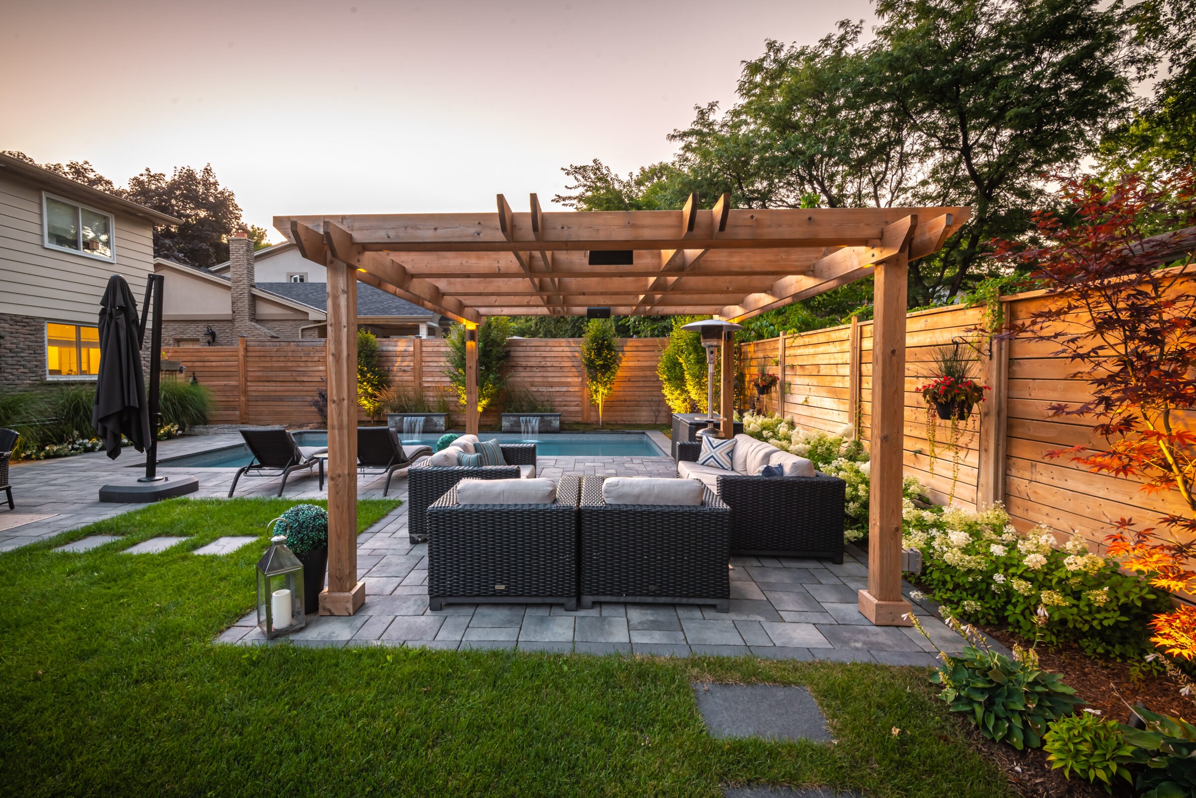 Backyard with pergola, patio furniture, and lush greenery, surrounded by wooden fence. Evening lighting enhances cozy atmosphere; well-maintained garden and plants.