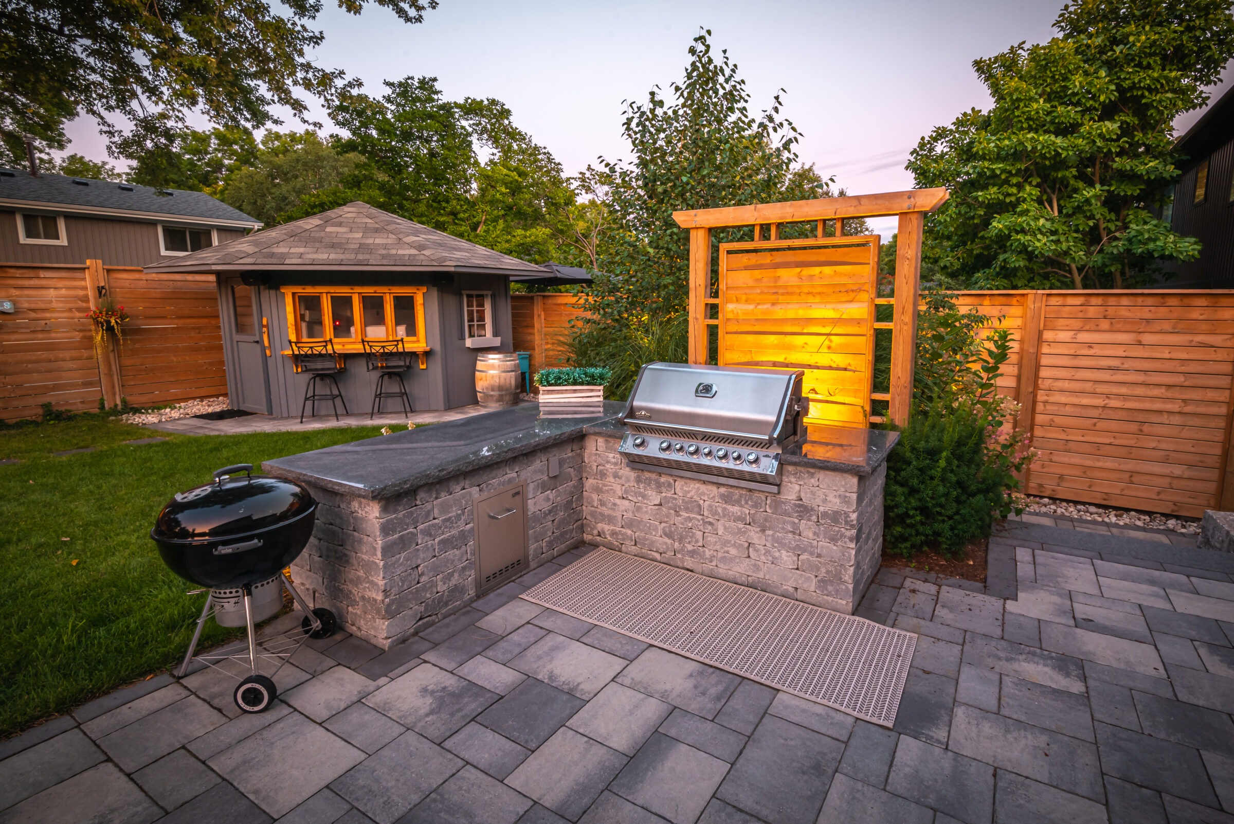A cozy backyard features a grill station, paver patio, and a small bar shed, surrounded by wooden fencing and lush greenery.