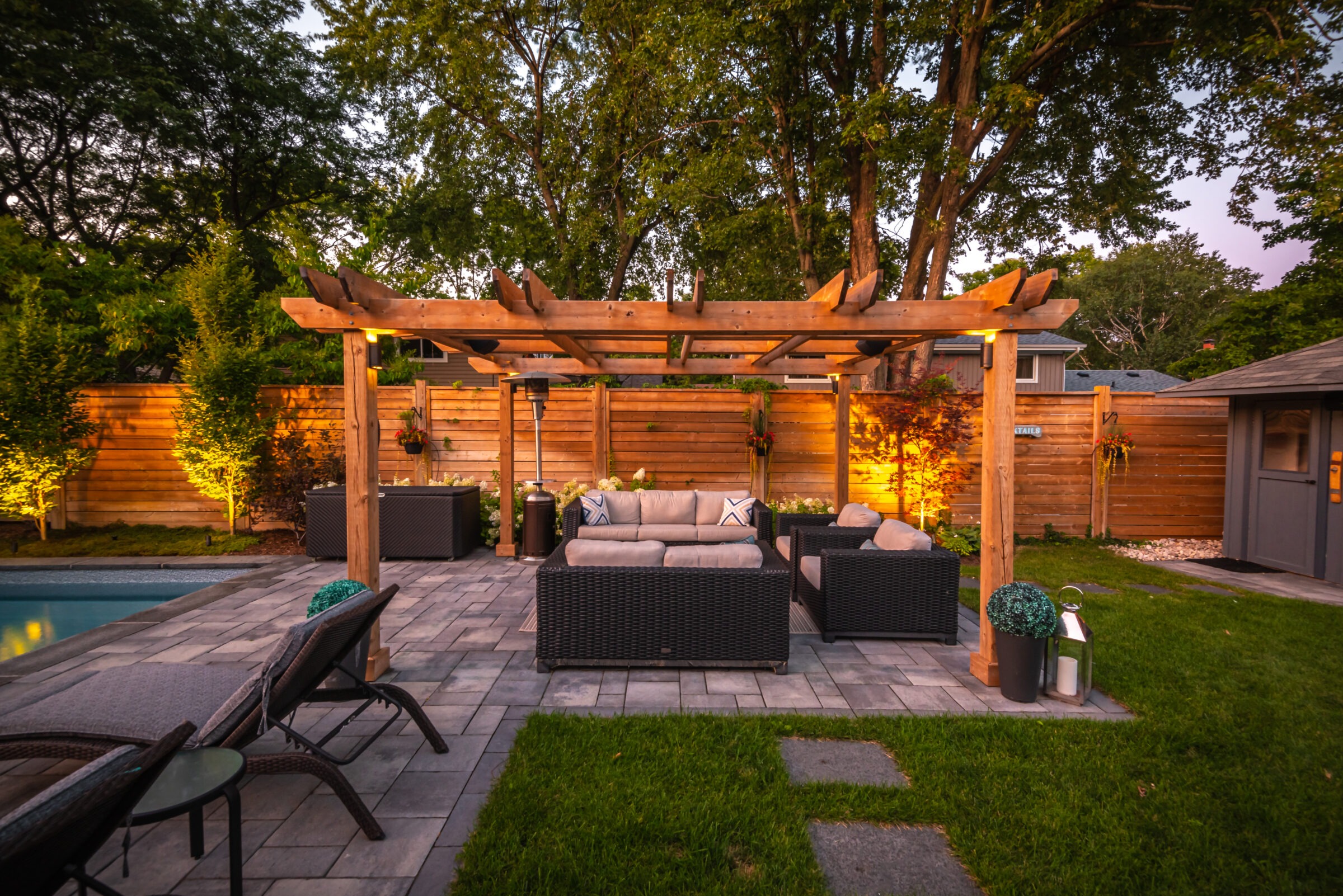 A backyard with wooden pergola, wicker furniture, and outdoor lighting, surrounded by trees and a wooden fence, near a pool.