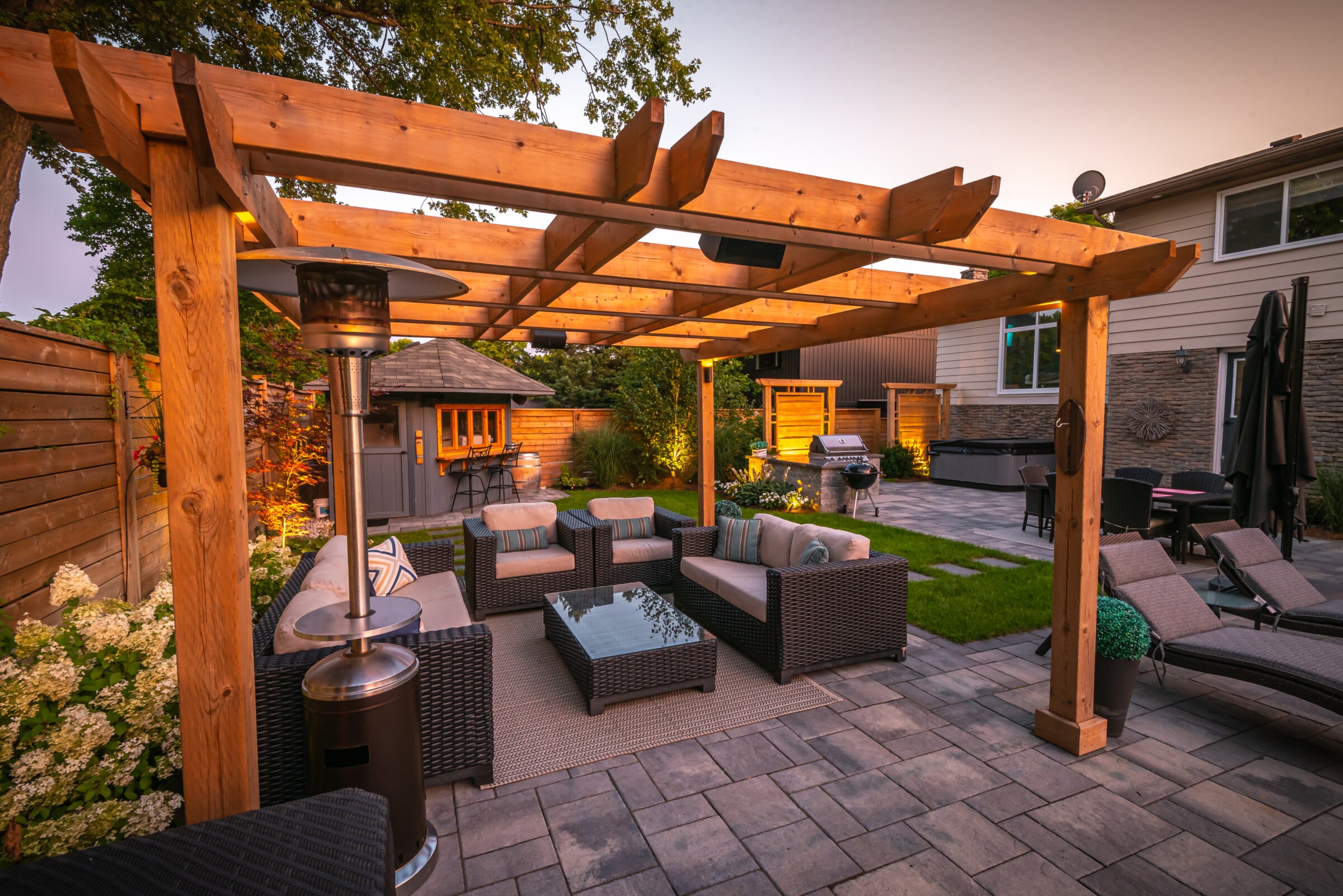 Wooden pergola over patio with wicker furniture, glass table, heater, and garden view; modern backyard setup with gazebo and barbecue grill.
