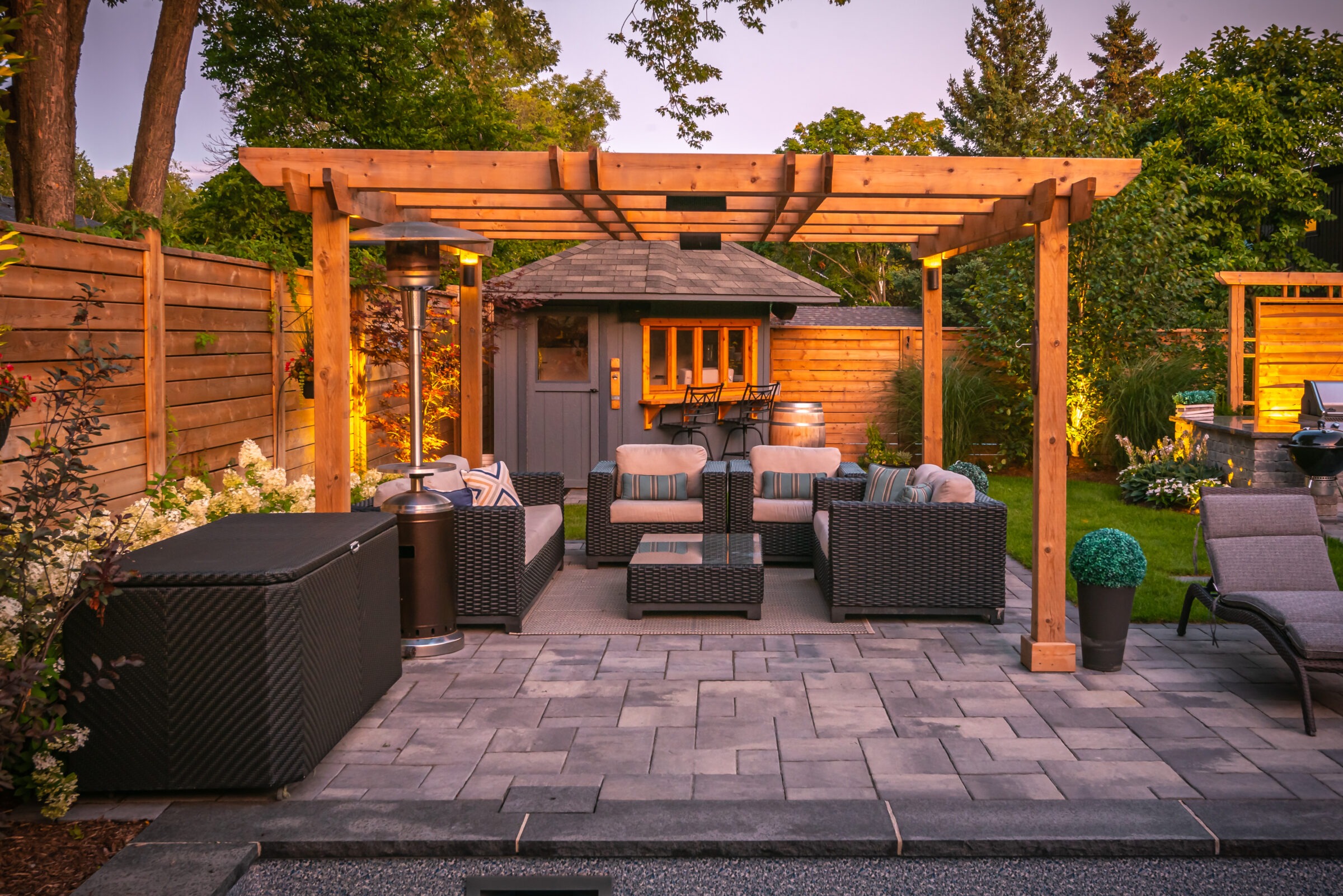 Cozy outdoor patio with wooden pergola, wicker furniture, and ambient lighting. Surrounded by lush greenery and wooden fencing in a serene garden setting.