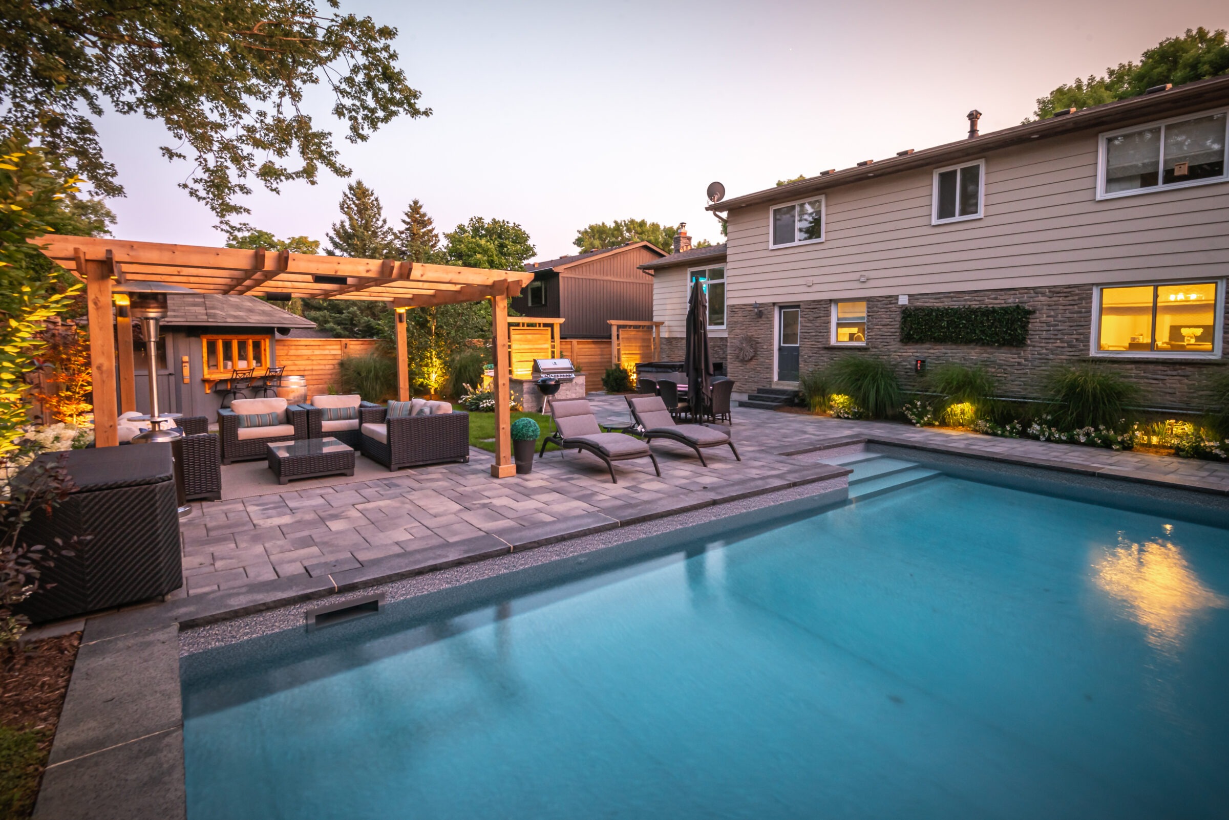 A cozy backyard with a swimming pool, pergola, outdoor seating, lounge chairs, and a house, surrounded by lush greenery at dusk.