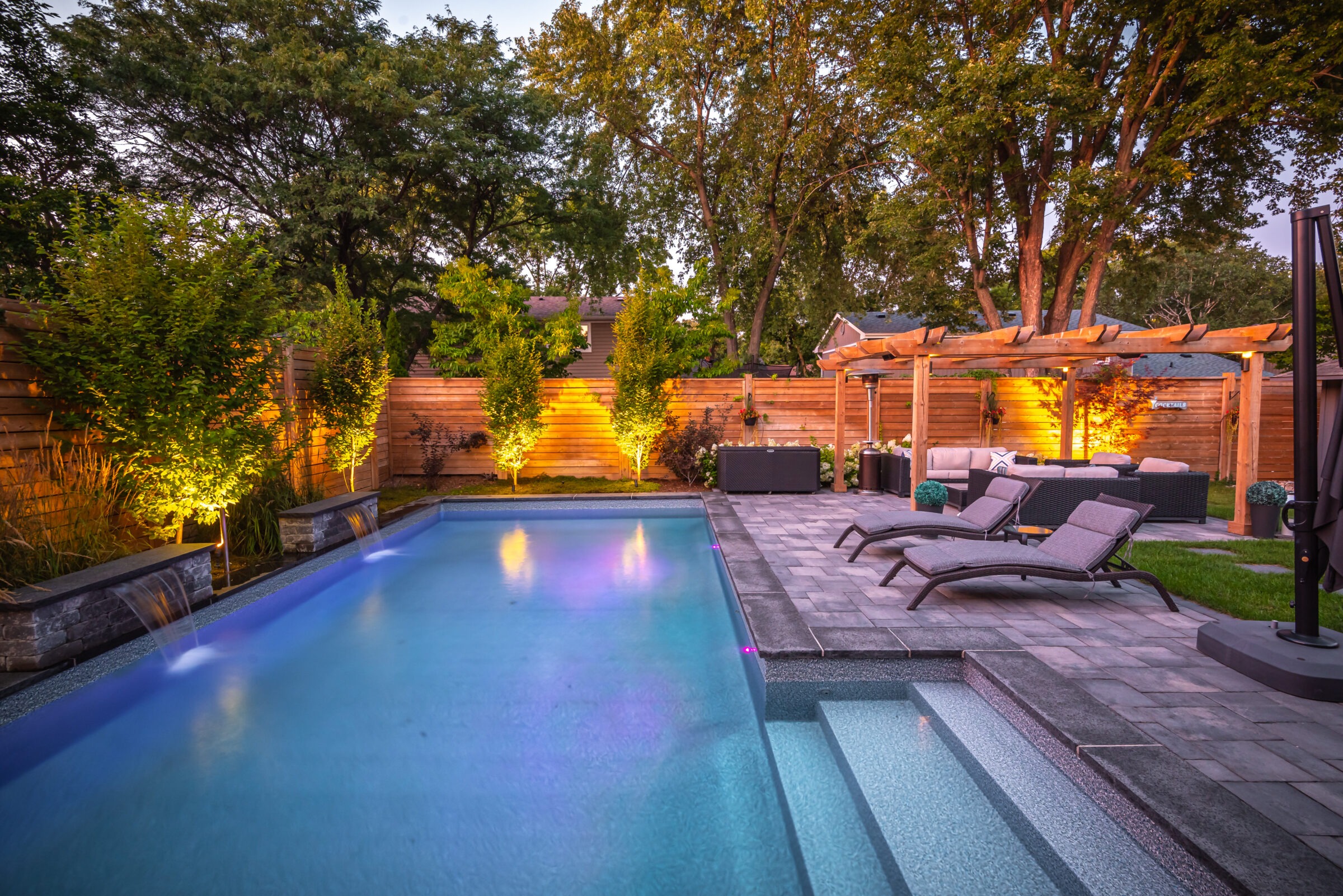 Backyard with illuminated pool, modern patio furniture, lounge chairs, pergola, and trees, creating a serene and inviting outdoor living space.