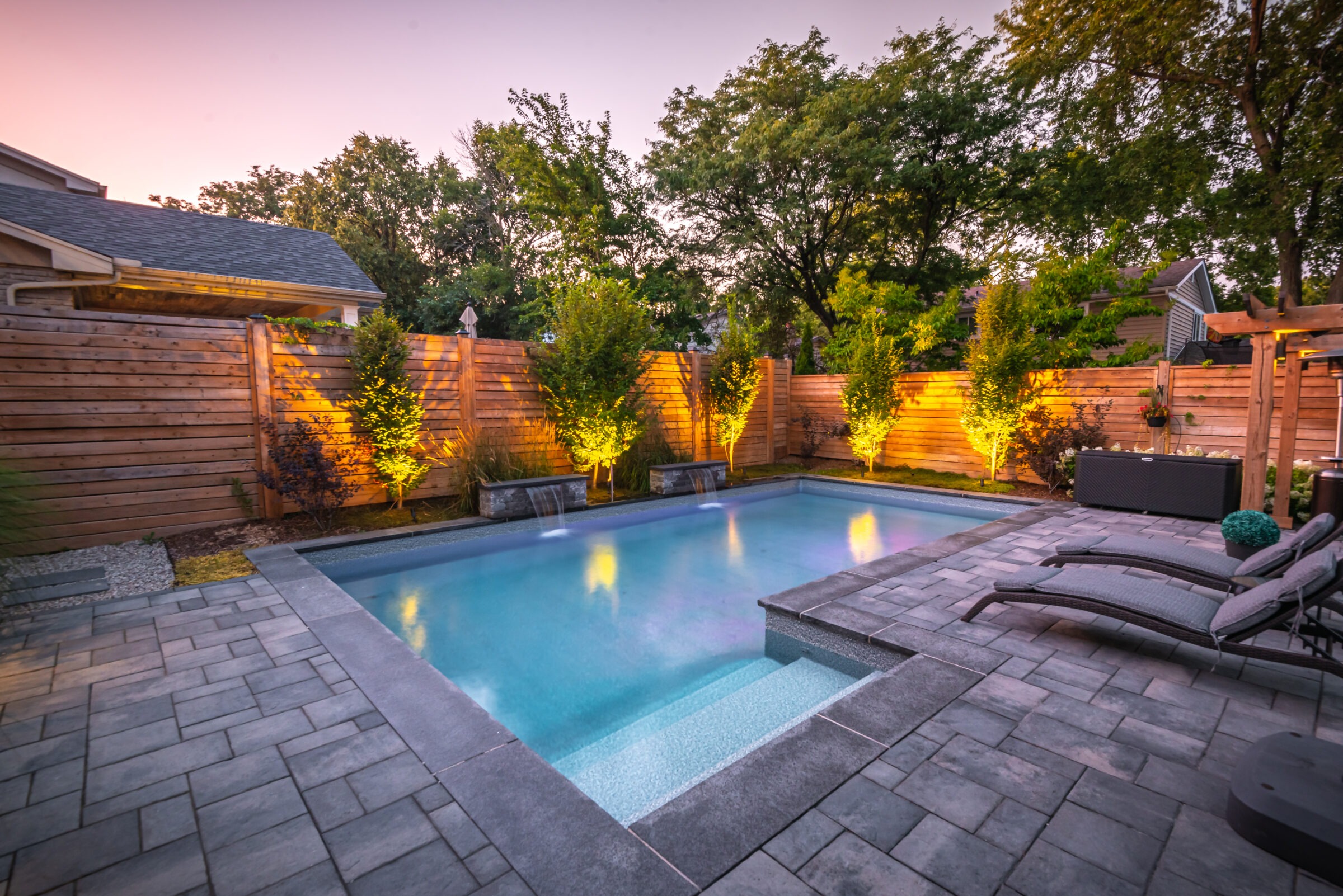 A serene backyard with a lit swimming pool, surrounded by greenery and wooden fencing, featuring stone seating and evening lighting.