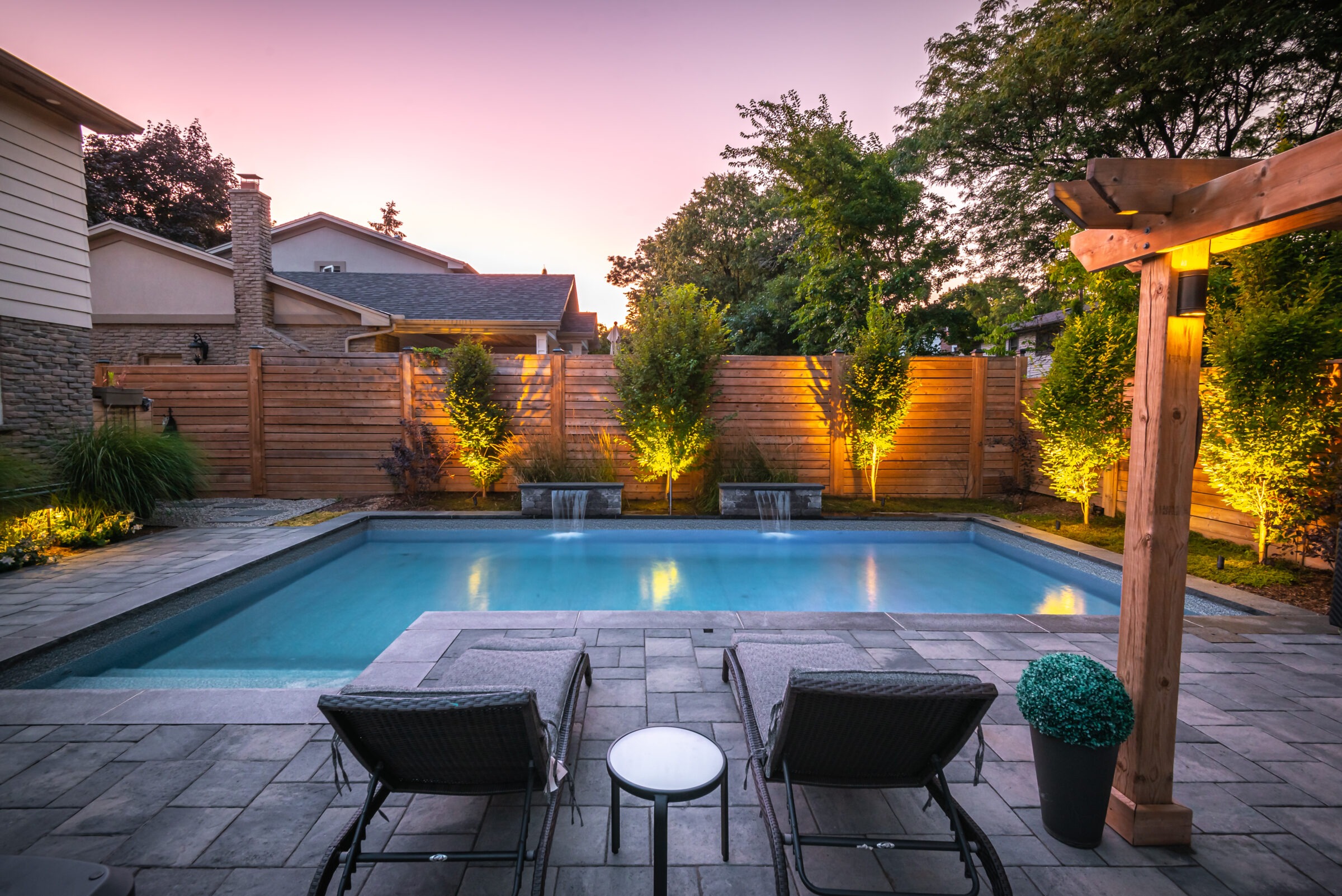 Backyard with a serene pool, illuminated trees, two lounge chairs, and a wooden pergola at dusk, surrounded by a wooden fence and houses.
